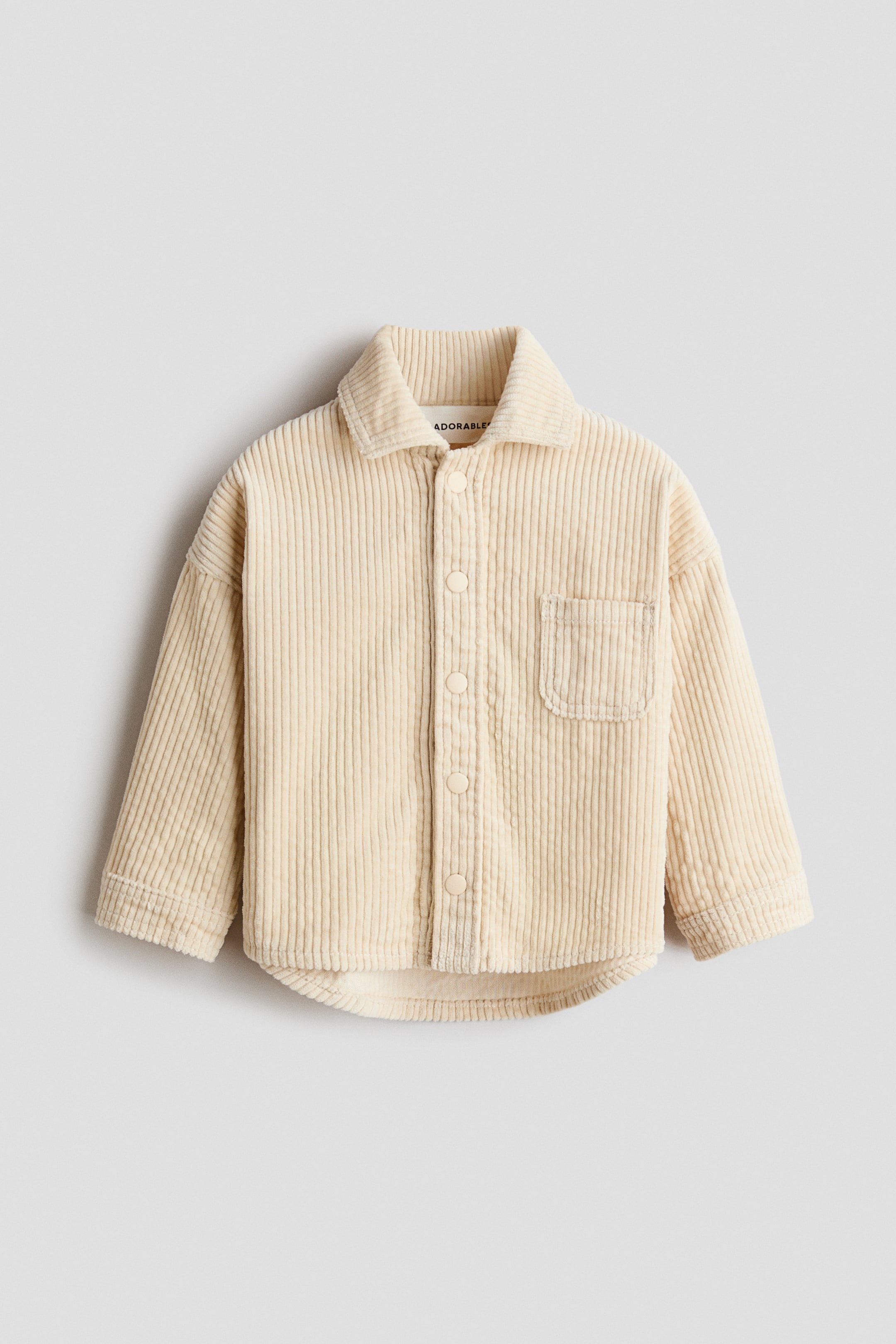 Agrandir l'image: Une chemise en velours côtelé crème est présentée de face, avec un col pointu et une patte de boutonnage complète ornée de boutons ronds assortis. Elle présente une seule poche plaquée sur la poitrine gauche, des manches longues et un ourlet arrondi.