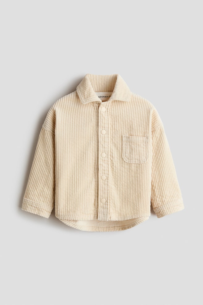 Se muestra una camisa de pana color crema de frente, con cuello en punta y una botonadura completa con botones redondos a juego. Presenta un solo bolsillo de parche en el pecho izquierdo, mangas largas y un dobladillo curvo. - 1