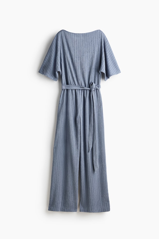 A blue jumpsuit featuring a ribbed texture throughout. It is designed with wide short sleeves and a boat neckline. A self-fabric tie belt cinches the waist and is tied in a knot, while the trousers flow into a wide-leg style. - 1