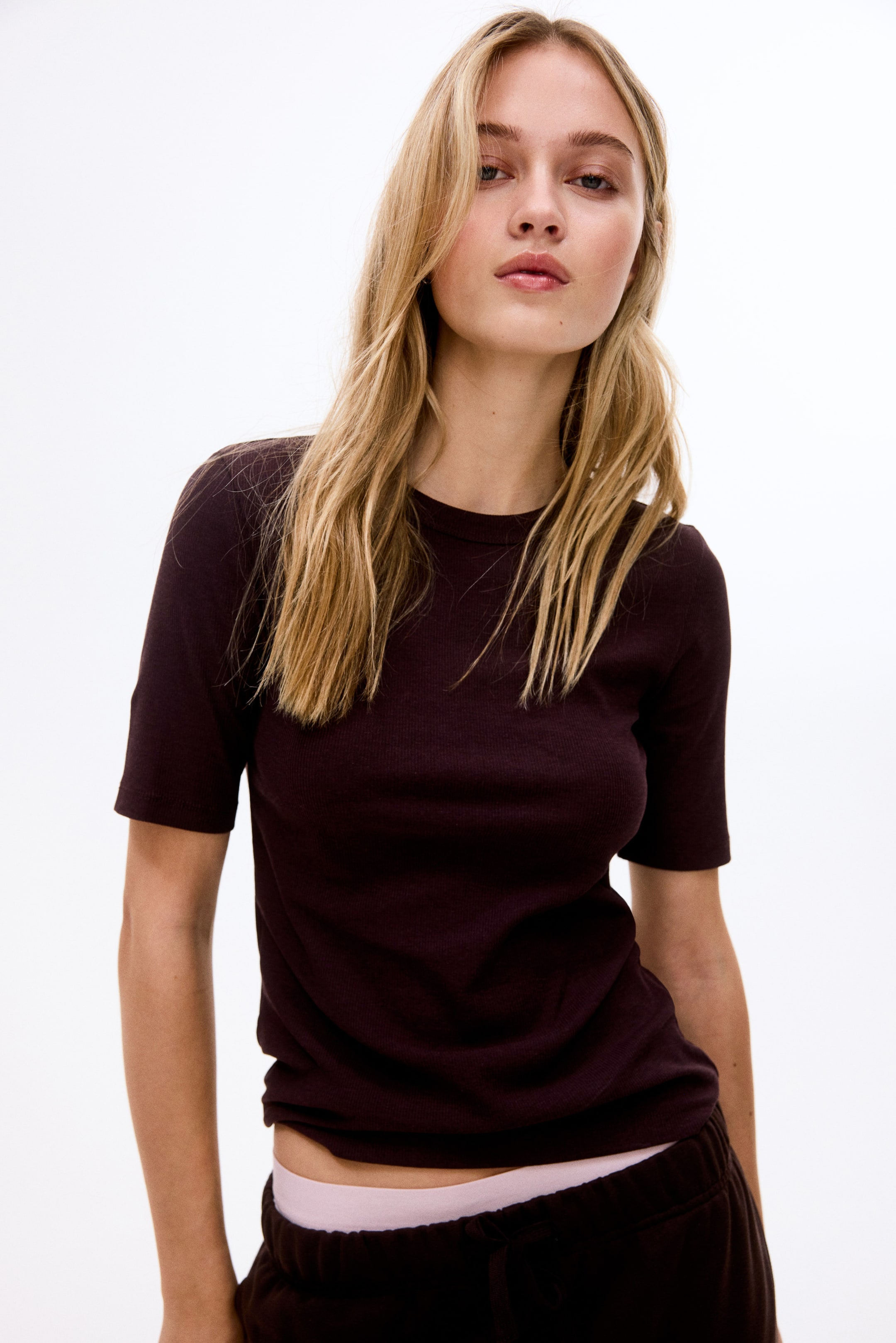 Se større billede: Den mørkebrune ribstrikkede t-shirt vender fremad og fremviser en tætsiddende silhuet med rund hals og korte ærmer. Dens kant interagerer med mørke bukser, der afslører en lyserød linning nedenunder.