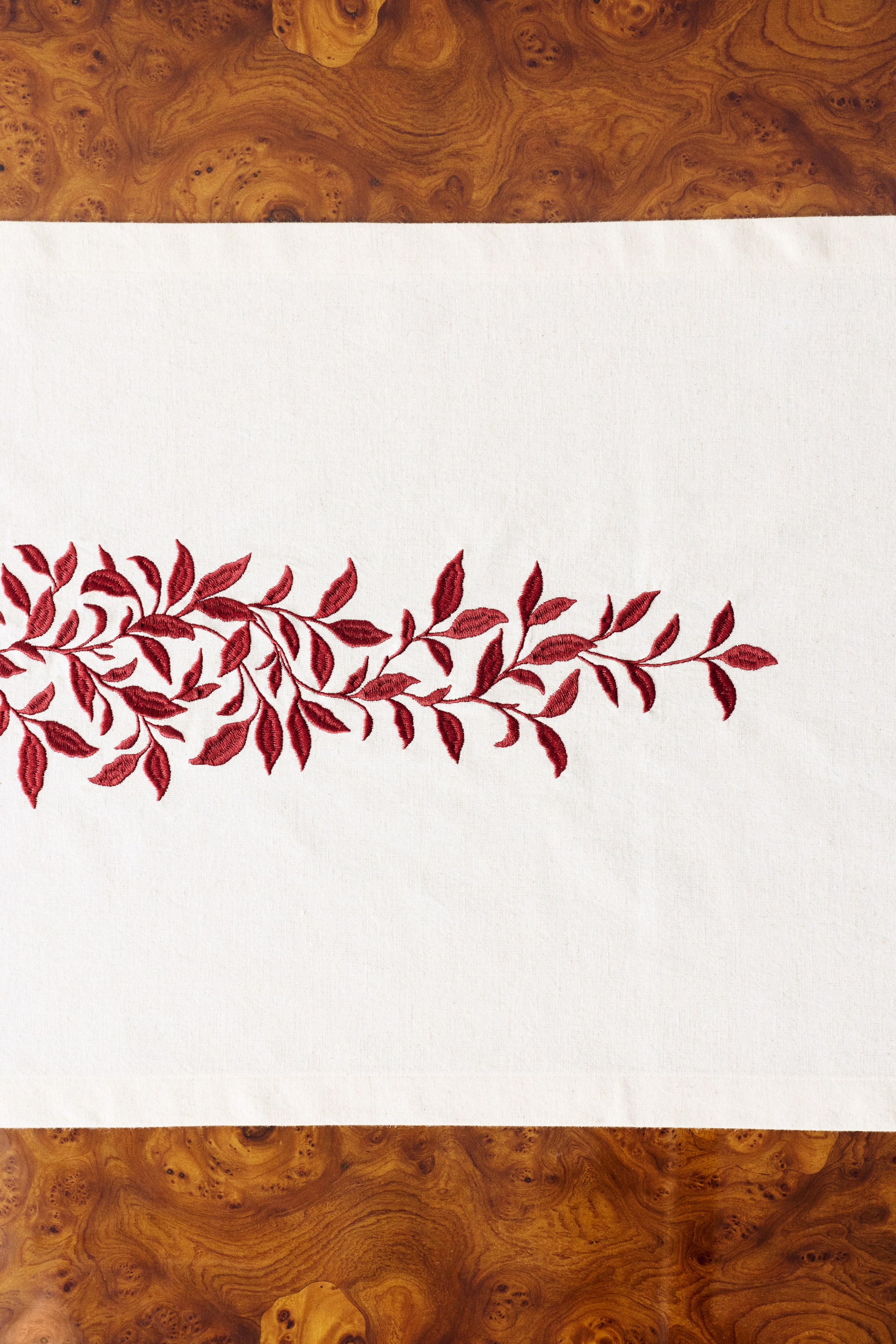 View larger image: An off-white cotton and linen weave table runner is shown from a top-down view, featuring a detailed, horizontal vine and leaf design embroidered in red satin stitching across its centre.