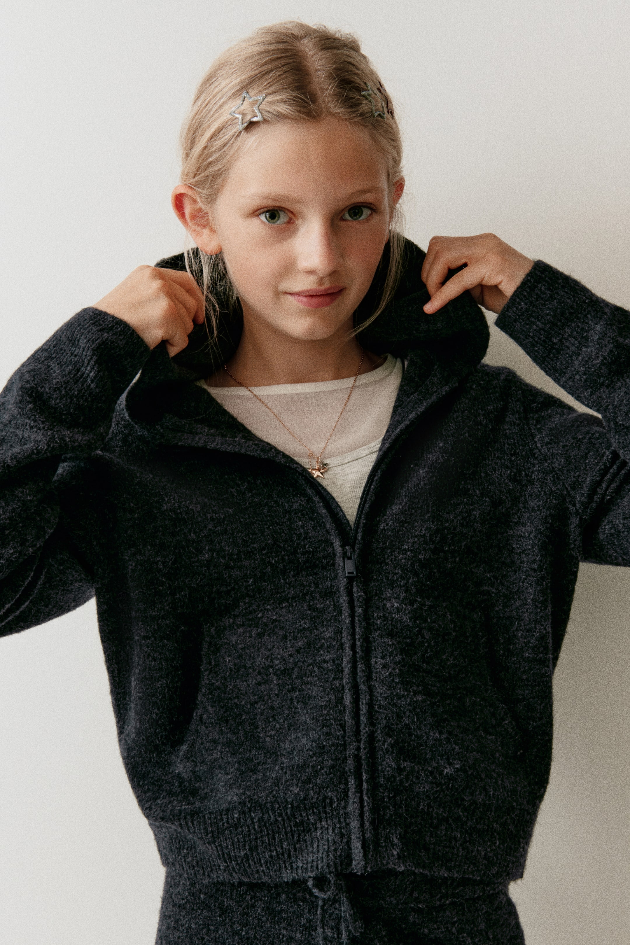 View larger image: A dark grey knit zip-up hoodie is worn facing front, showcasing its soft, lightly brushed texture and a central front zipper. The wearer is holding the hood, highlighting the ribbed hem, ribbed cuffs, and relaxed fit of the garment layered over a light-coloured top.