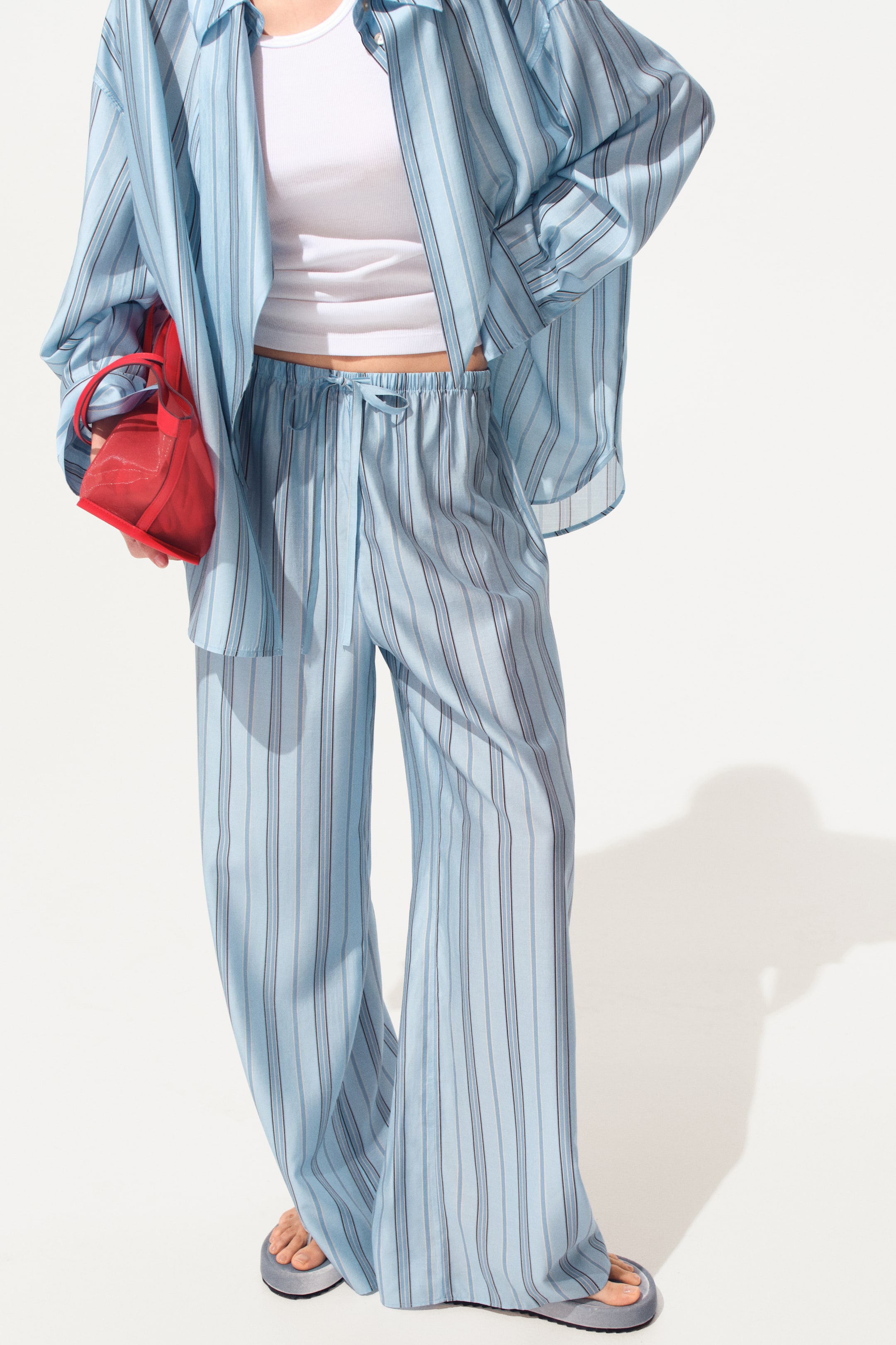 View larger image: The model is wearing loose-fit pants in a light blue striped woven fabric, featuring a high, elastic waistband with a narrow drawstring and wide legs. A matching striped shirt is layered over a white top.