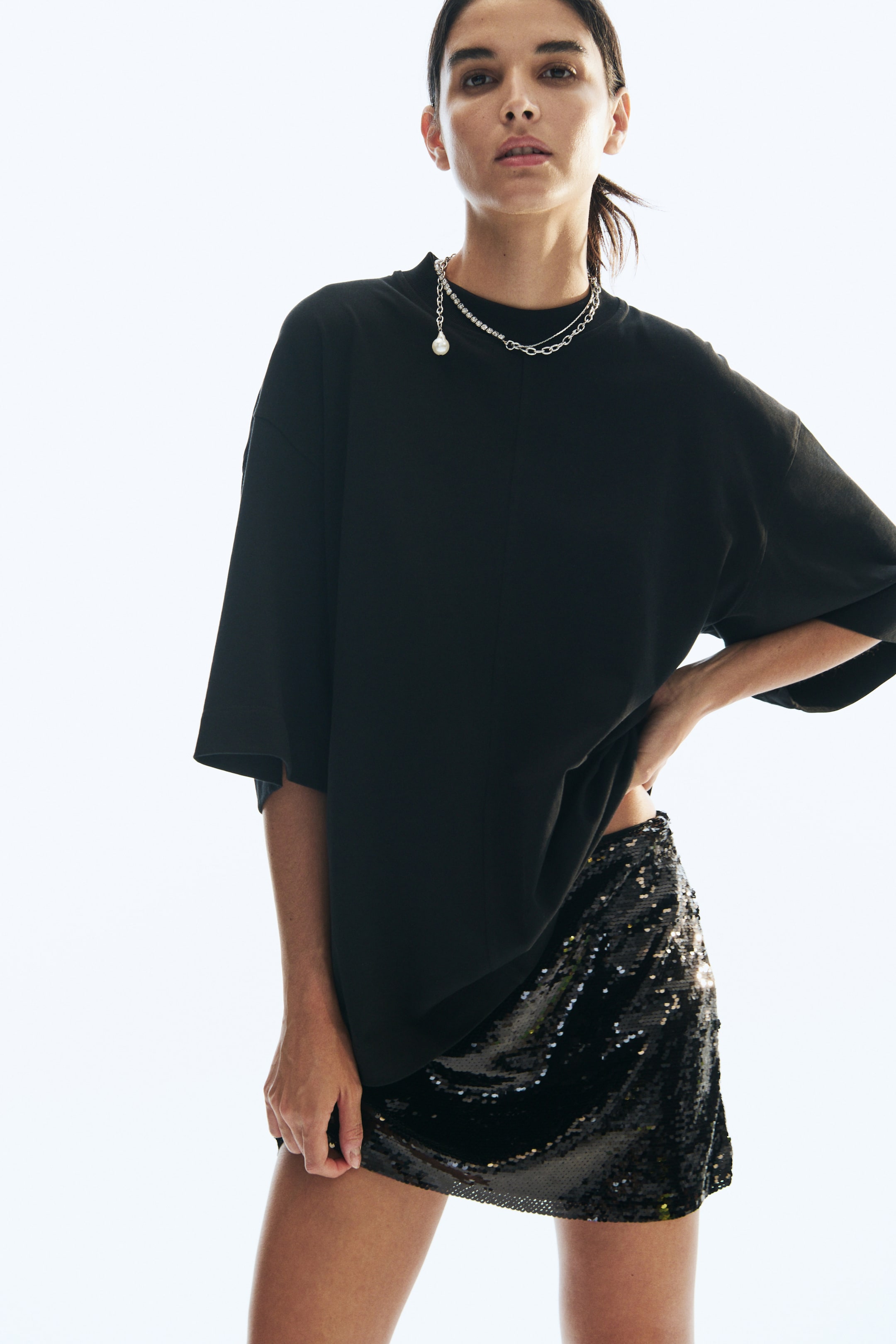 View larger image: A sequinned mesh mini skirt is being worn by the model, paired with an oversized black T-shirt. The model is holding the raw-edge hem of the skirt with one hand while resting the other hand on their hip, slightly lifting the T-shirt to reveal the skirt’s waistband.