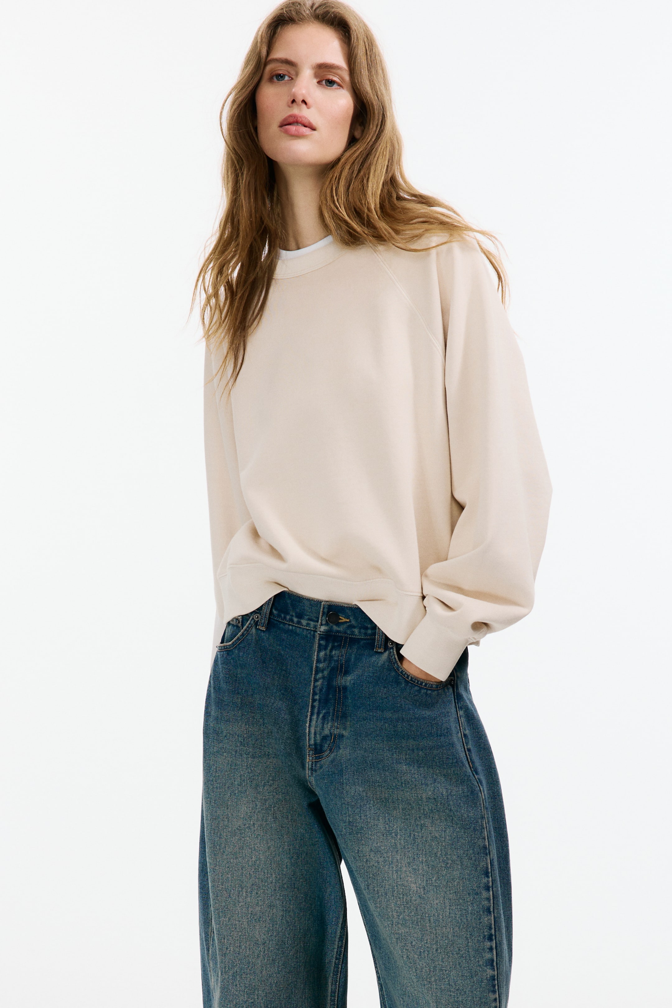 View larger image: The beige sweatshirt showcases a crew neck with a visible white t-shirt layered underneath, raglan sleeves, ribbed cuffs, and a ribbed hem, offering a relaxed fit over blue jeans.