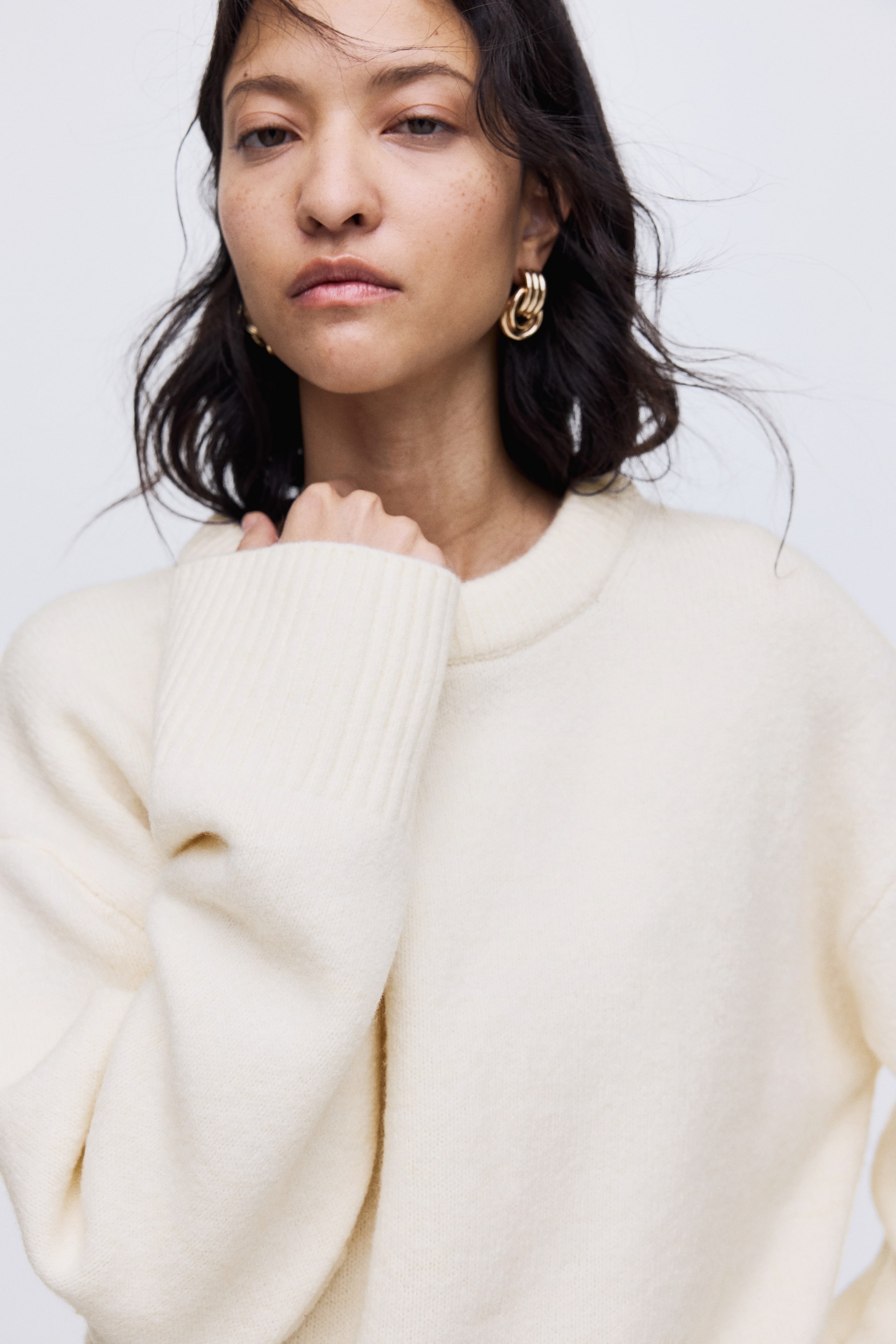 View larger image: A cream-colored crew neck sweater is being presented from a front-right angle, featuring a soft, slightly fuzzy knit texture and wide, ribbed cuffs on its sleeves.