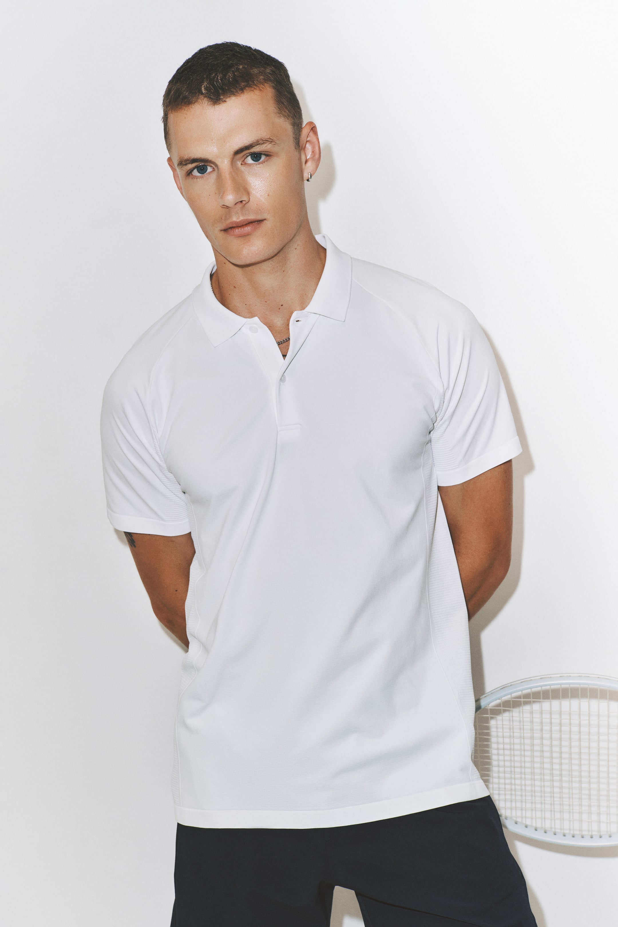 Grotere afbeelding bekijken: Een witte Regular Fit Tennis T-shirt met DryMove? wordt gedragen door het model. Het heeft een polokraag, een sluiting met drie knopen, raglanmouwen en een subtiel gestructureerd patroon aan de zijkanten en manchetten. Het model combineert het met zwarte shorts.