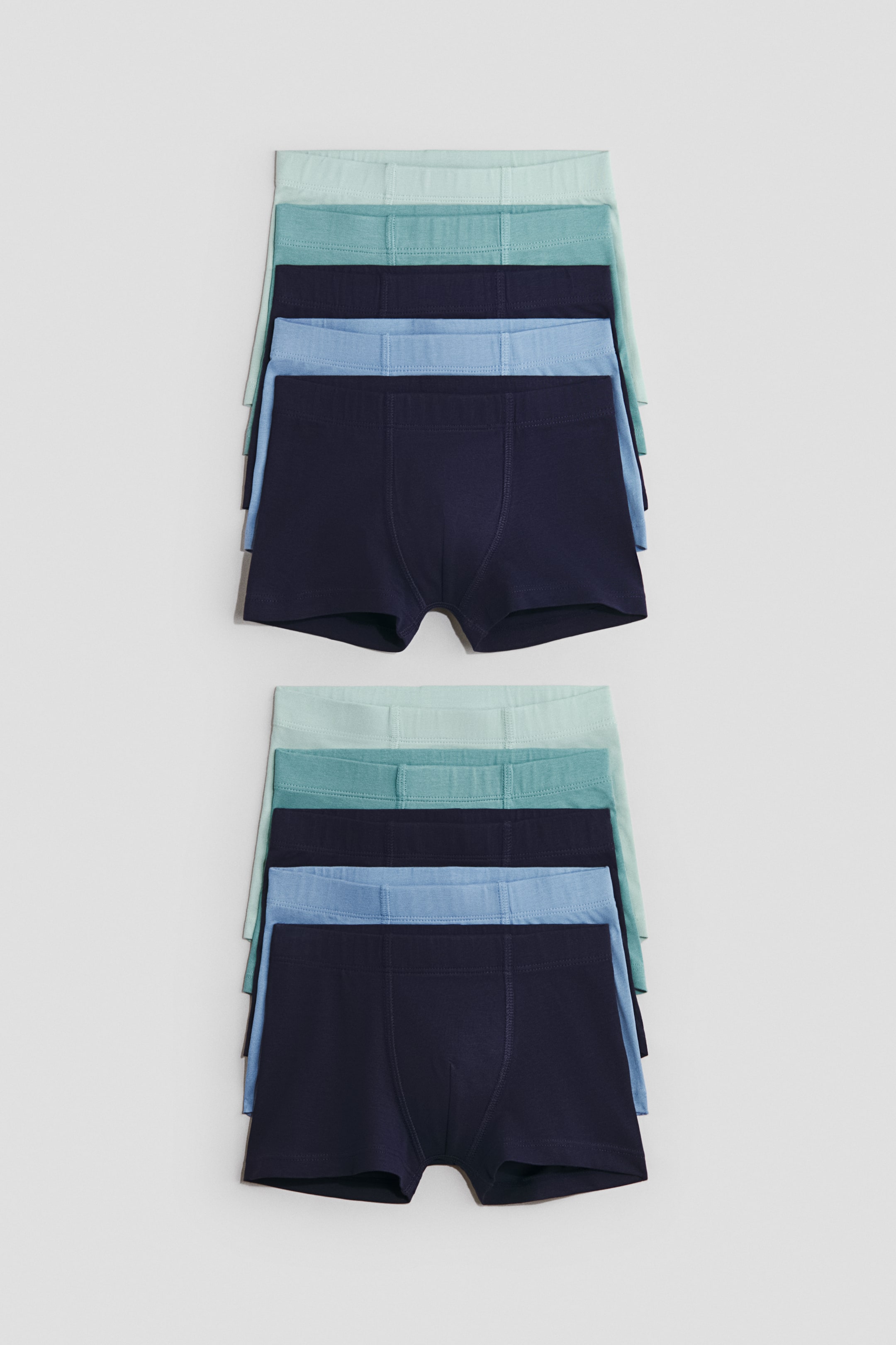 View larger image: Trunks are displayed in two neatly stacked groups, each featuring five pairs with short legs, an elasticated waist, and a lined front. These soft cotton jersey trunks showcase a range of blue and green shades, with the darkest blue pair fully visible at the bottom of each stack, and are lying flat, viewed from above.