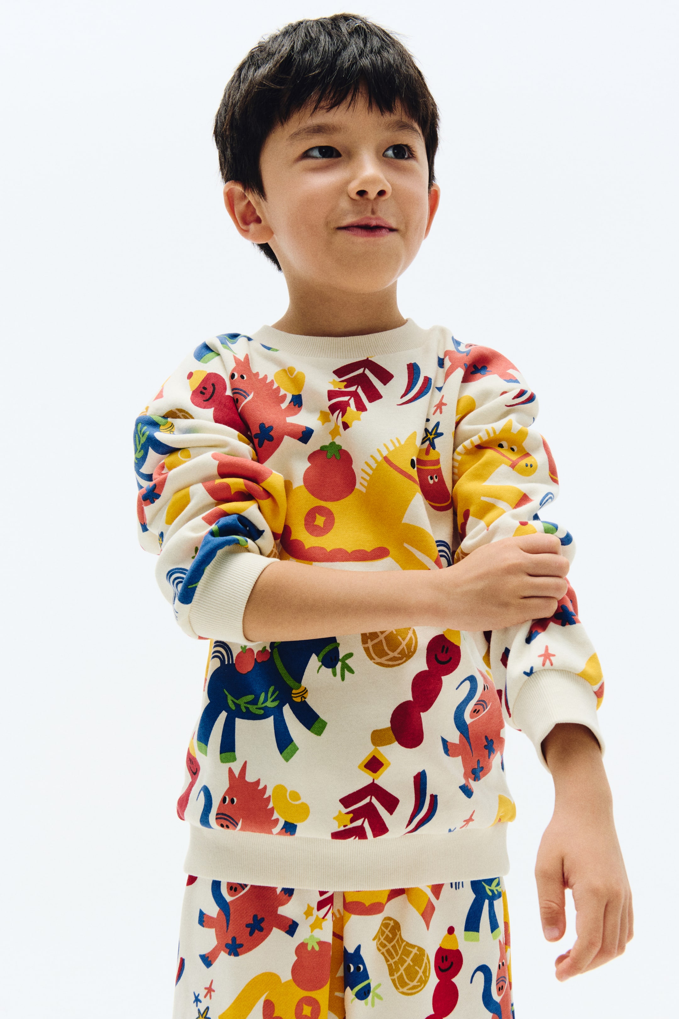 View larger image: A cream sweatshirt and matching pants for kids feature a vibrant all-over print of stylised horses, peanuts, and abstract shapes in red, yellow, and blue. The sweatshirt comes with a crew neck, slightly puffed long sleeves with ribbed cuffs, and a ribbed hem.