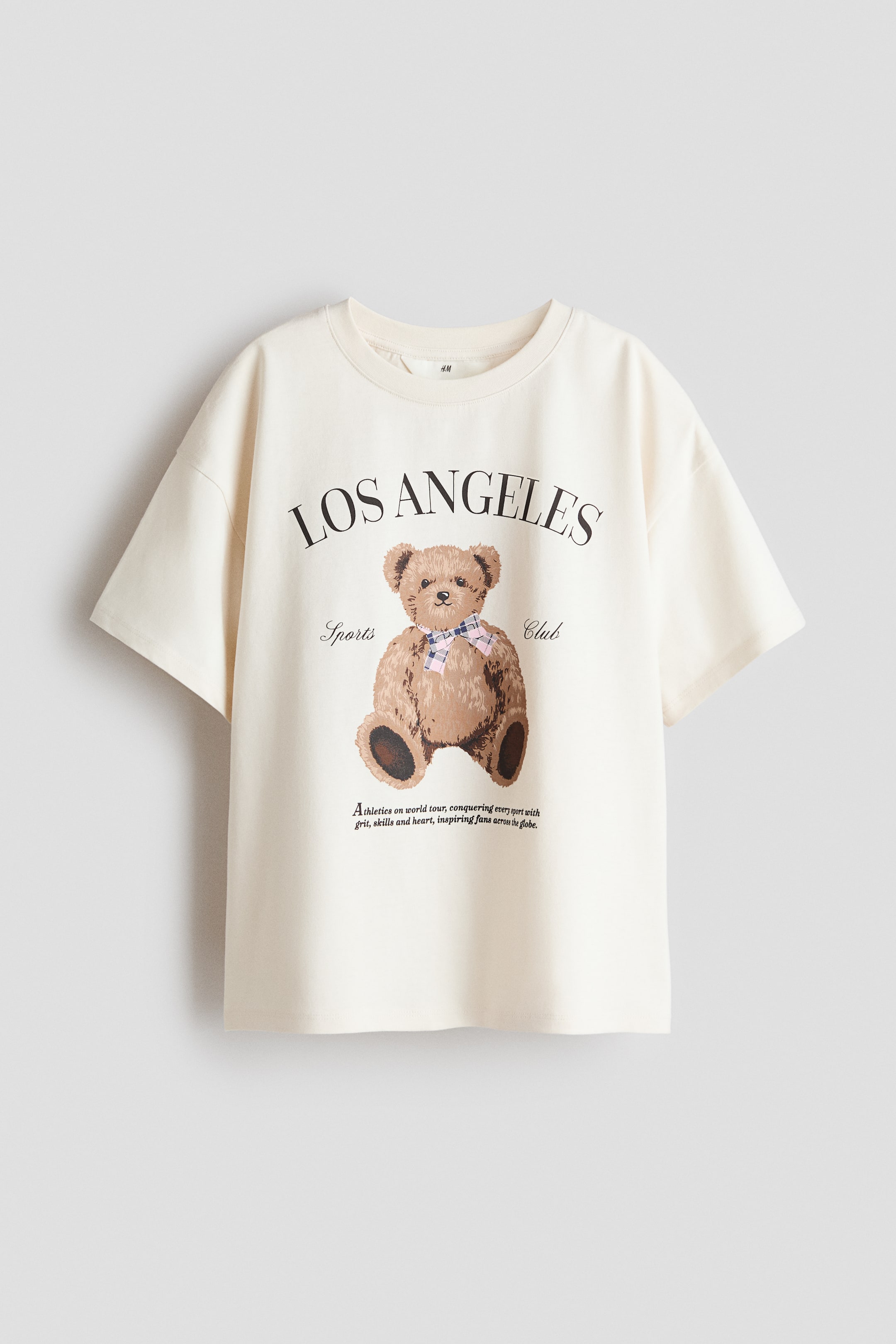 View larger image: An oversized t-shirt crafted from soft, cream-coloured cotton jersey, featuring a round, rib-trimmed neckline, dropped shoulders, and a straight-cut hem. The front showcases an arched "LOS ANGELES" text print above a brown teddy bear graphic adorned with a blue and pink plaid bow tie, accompanied by "Sports Club" text on the sides and smaller descriptive text below.