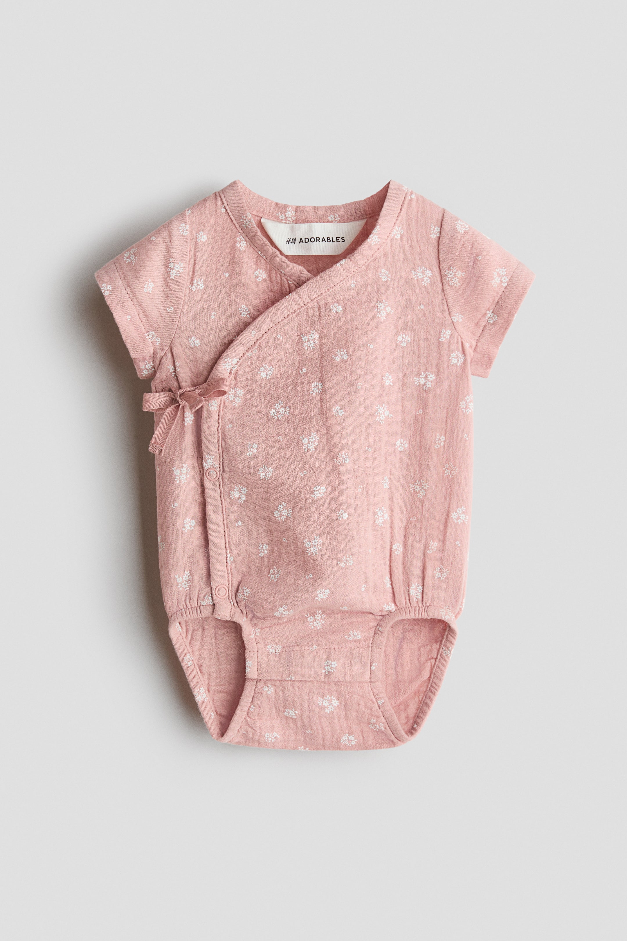 Grösseres Bild ansehen: Ein rosa Baby-Strampler zeigt eine geknitterte Stoffstruktur mit einem weißen Blumenmuster. Er verfügt über kurze Ärmel, ein Wickeldesign, das auf der linken Seite mit einer Schleife gebunden wird, und Druckknöpfe im Schritt.