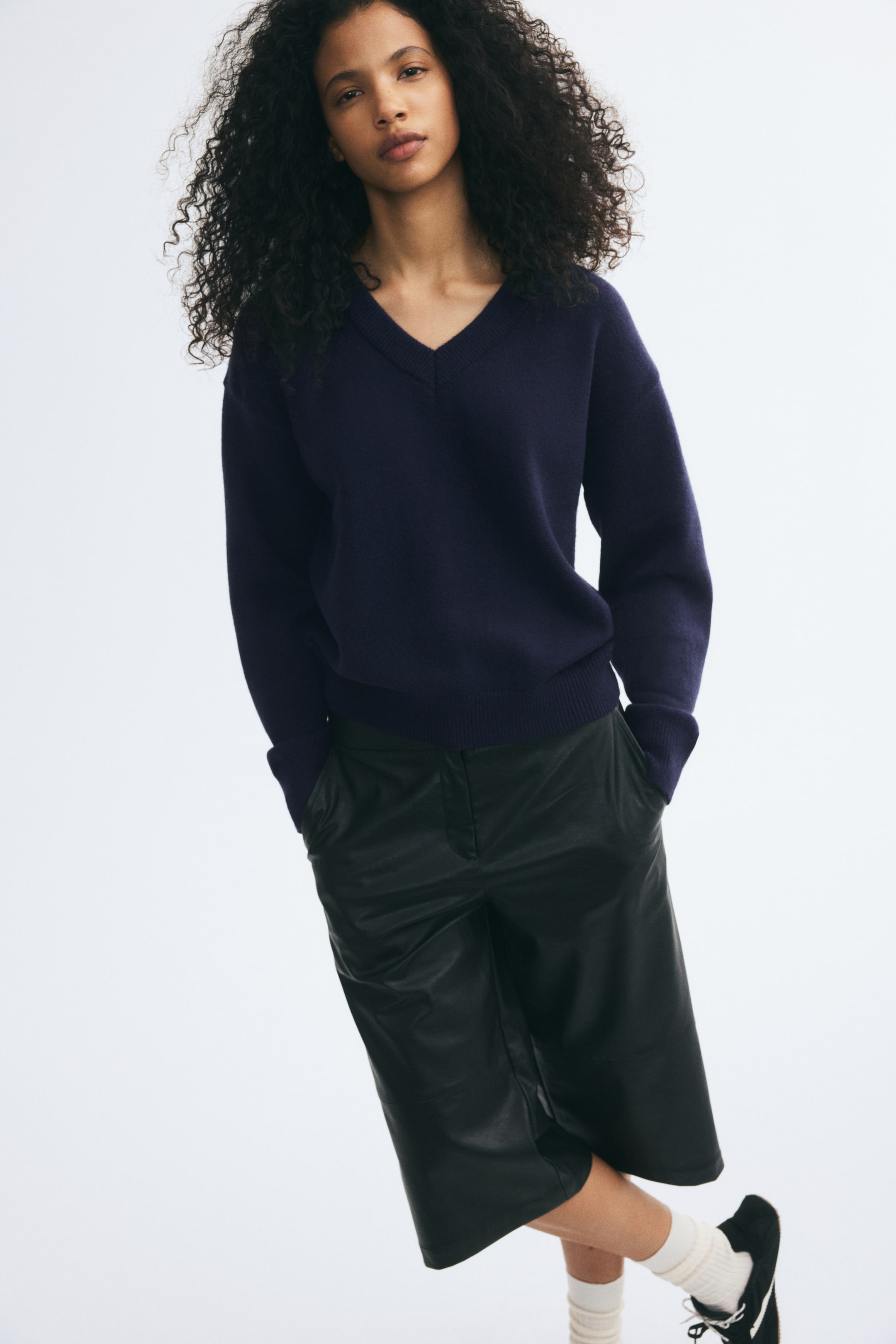 Agrandir l'image: Un pull en maille bleu foncé, doté d’un col en V côtelé, d’épaules tombantes ainsi que de poignets et d’un ourlet côtelés, est porté par le mannequin. Le pull est associé à un short large effet cuir noir, à des chaussettes blanches et à des baskets noires.