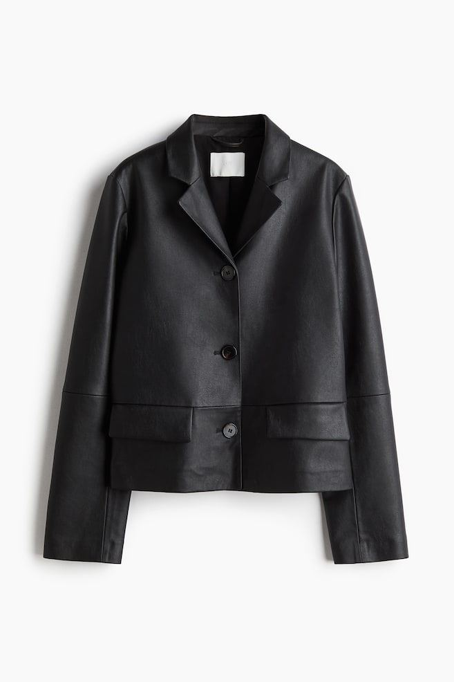 Une veste noire présente son DEVANT, avec une surface lisse et légèrement texturée. Elle est conçue avec des revers crantés, trois boutons ronds et sombres sur le devant, et deux poches à rabat rectangulaires sont positionnées à l'avant inférieur. - 1