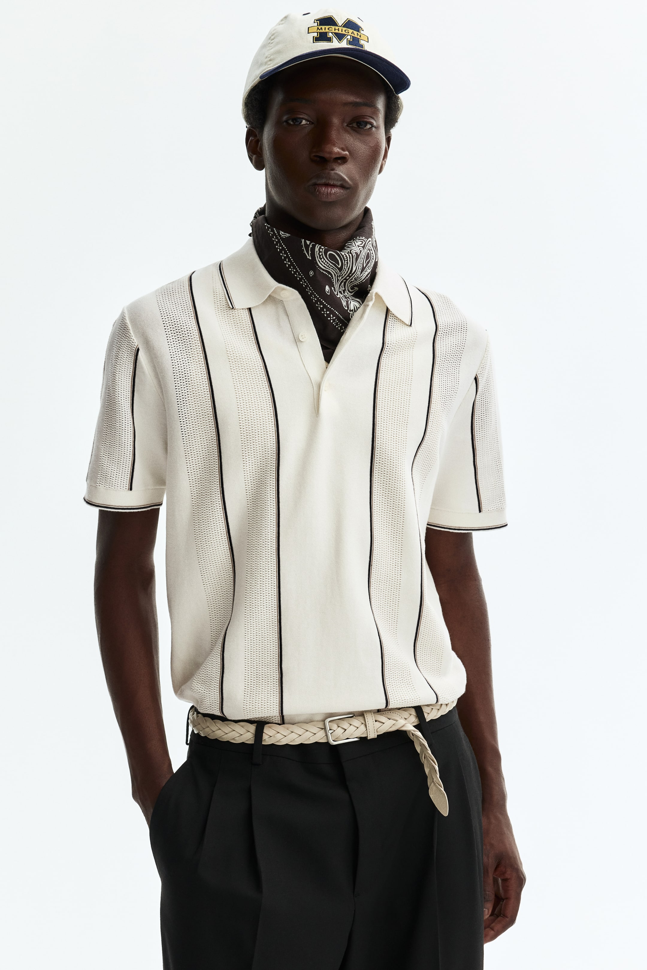 Ampliar la imagen: La camisa polo de punto crema presenta rayas verticales, alternando entre paneles sólidos y perforados, un cuello polo, una tapeta de dos botones y mangas cortas acanaladas con un ribete oscuro. Un pañuelo oscuro con patrón está metido bajo su cuello, y la camisa se muestra metida en pantalones negros, asegurada por un cinturón ligero trenzado.
