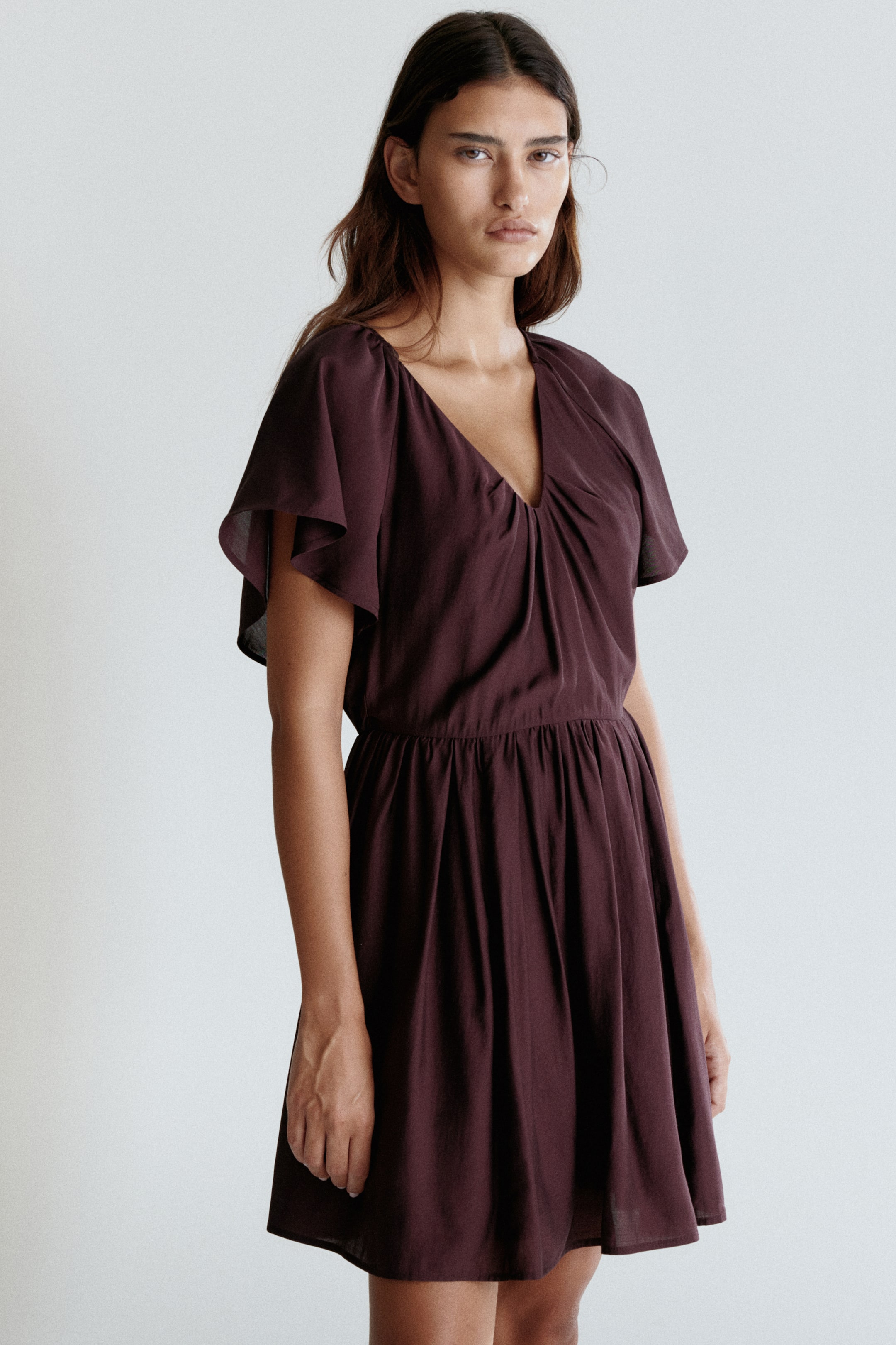 View larger image: A dark plum dress is presented from a front view, slightly angled to the right. It is featuring a V-neckline with central gathering on the bodice, short flutter sleeves, and an elasticated waist that leads into a gathered A-line skirt.