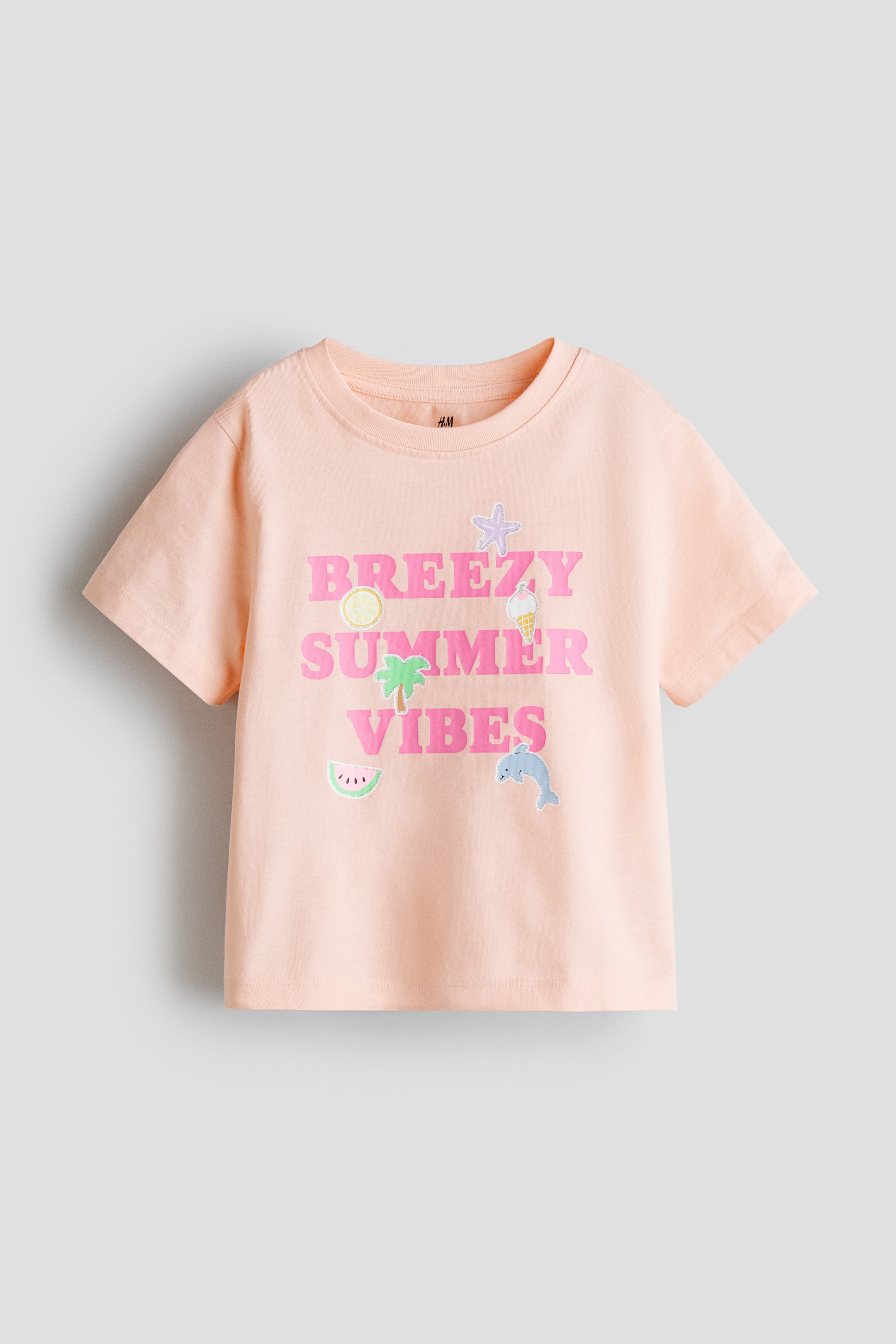 View larger image: A front view of a light peach t-shirt featuring a ribbed trim around the neckline. The t-shirt displays "BREEZY SUMMER VIBES" in pink capital letters, accompanied by embroidered summer-themed icons including a starfish, lemon, ice cream, palm tree, watermelon, and dolphin.