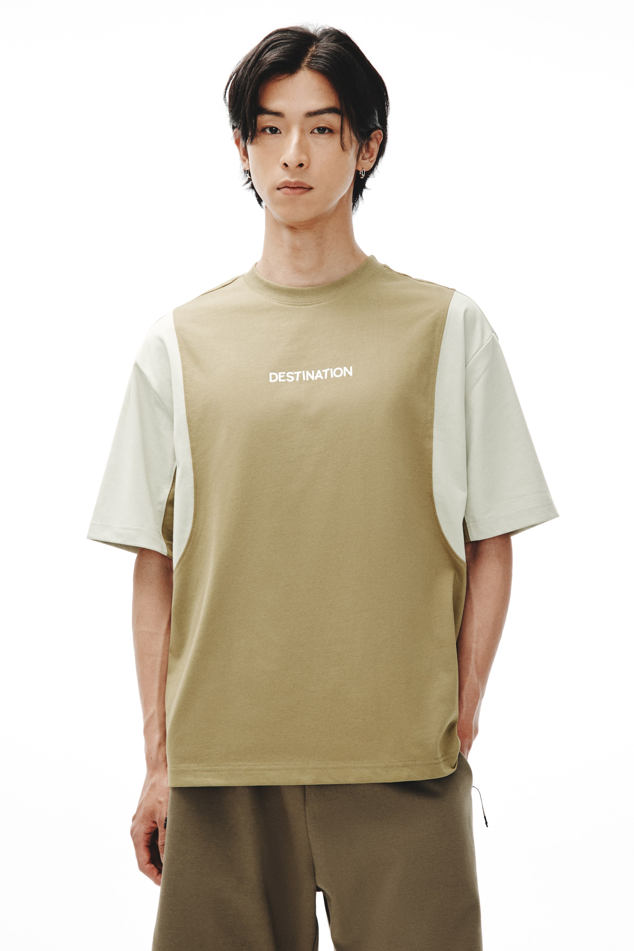 拡大画像を表示: モデルが着用しているのは、ツートーンのクルーネックTシャツです。カーキ色のメインボディに、胸元には「DESTINATION」という白い文字がプリントされています。また、より明るいベージュのカラーブロックサイドパネルが短い袖まで伸びています。