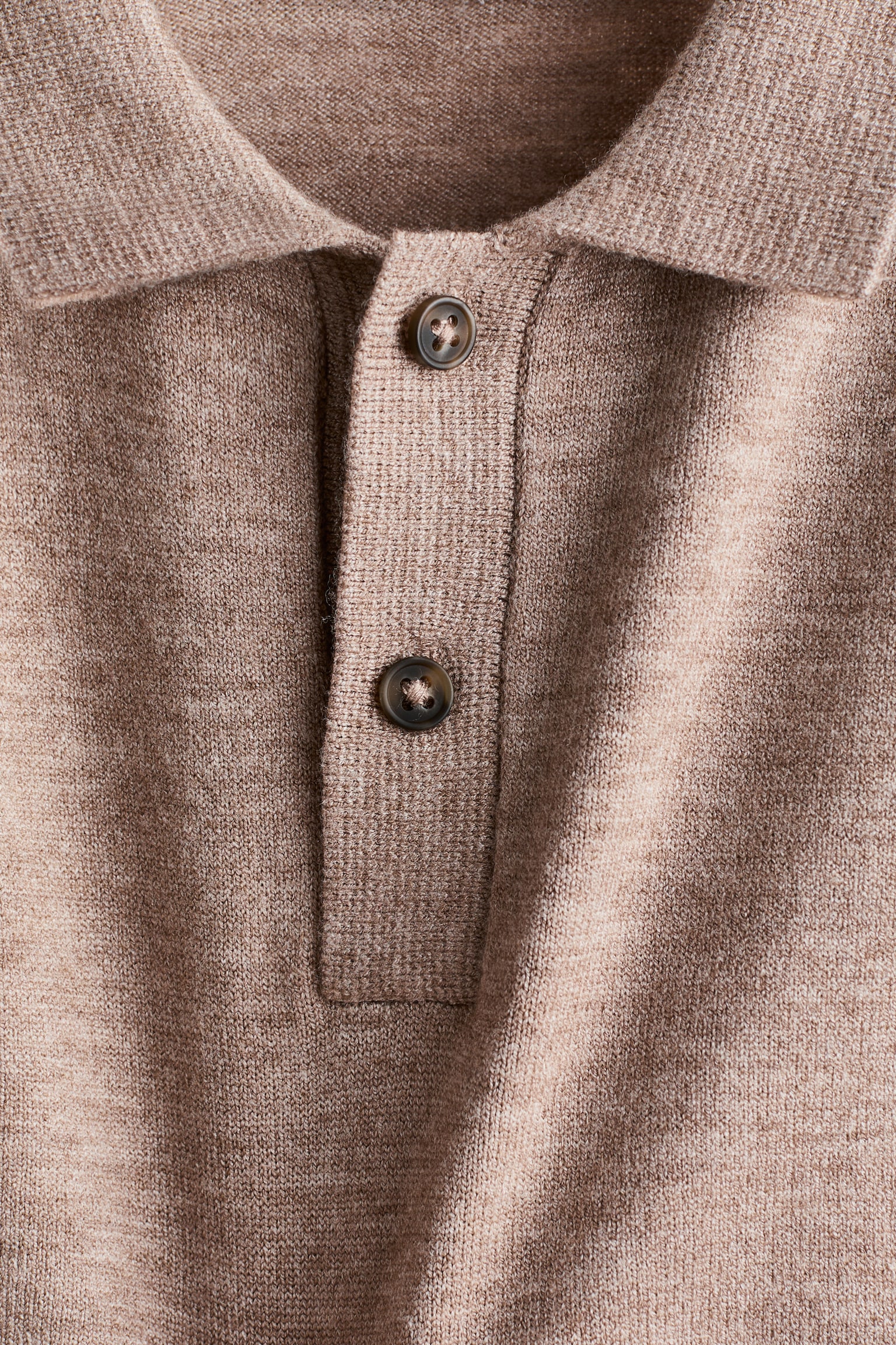 A long-sleeve rugby shirt in a soft, fine knit is shown in a close-up front view of its collar and button placket. The shirt features two brown buttons and a heathered light brown knit texture.
