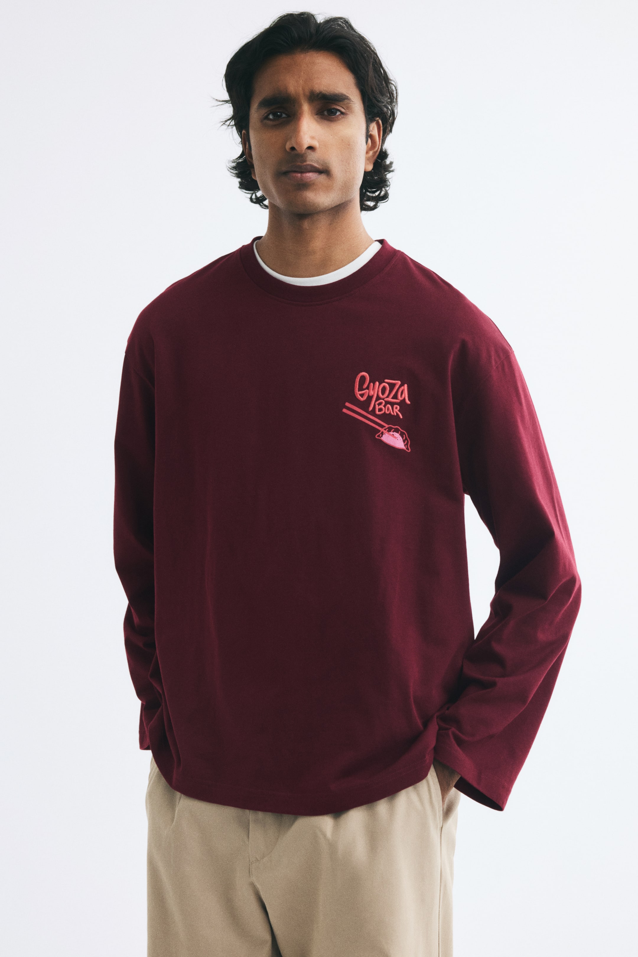 View larger image: A burgundy long-sleeve T-shirt featuring a ribbed crew neckline, dropped shoulders, and a small graphic print on the left chest is being worn by the model. The relaxed-fit tee is layered over a white undershirt and styled with beige trousers.