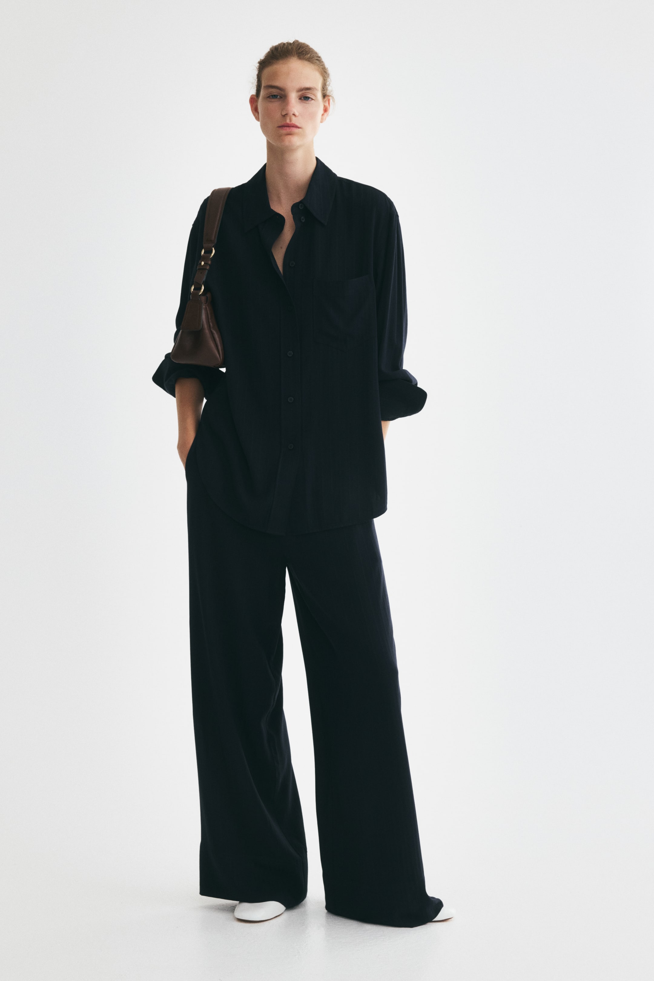View larger image: A dark blue co-ord set, featuring a long-sleeved shirt and wide-leg pants, is being worn by the model. The shirt has a classic collar, button-front closure, a chest pocket, and rolled-up cuffs, with some buttons unfastened at the neckline. The model is placing her hands into the pants' pockets, carrying a brown shoulder bag on her shoulder, and wearing white flats.