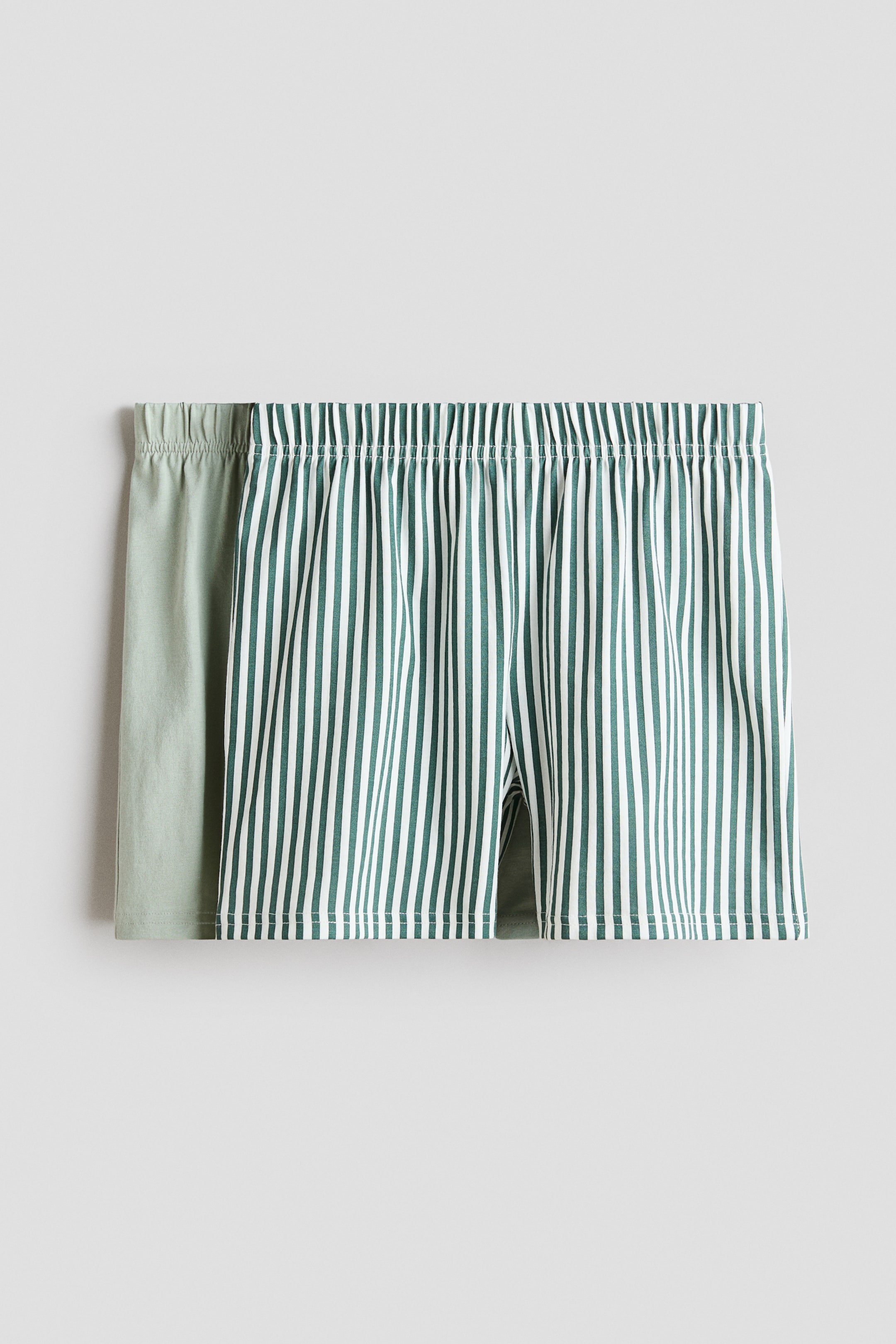 View larger image: Two pairs of pyjama shorts in soft cotton jersey are presented from the front, featuring elasticated waists. The front pair displays vertical dark green and white stripes, while the partially visible back pair shows a solid light green colour.