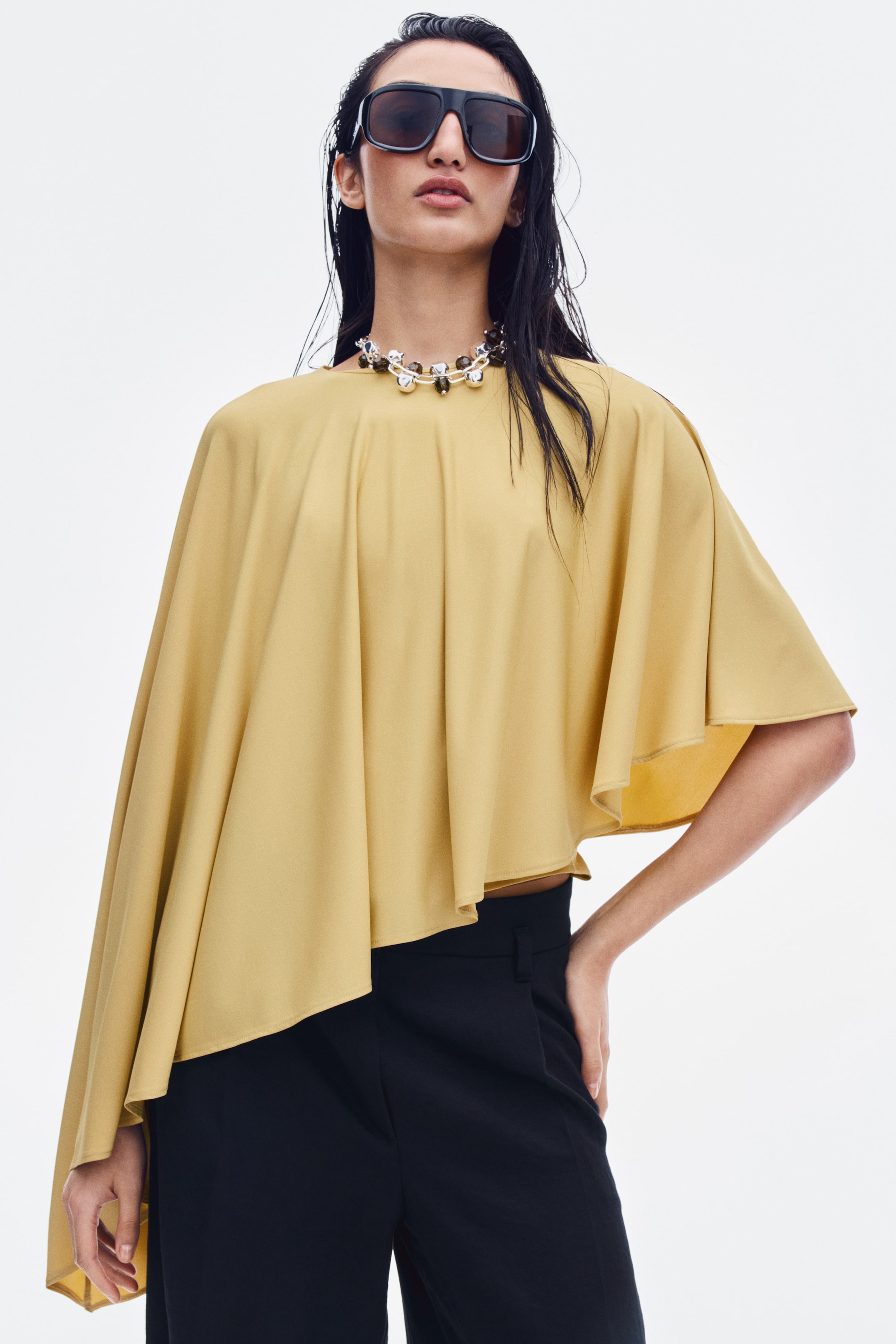 View larger image: A mustard-yellow sleeveless bodysuit with a boat neck is being worn by the model, featuring a draped, cape-like overlay with a dramatic asymmetrical hem. The bodysuit is tucked into high-waisted black trousers and styled with a chunky beaded necklace and oversized sunglasses.