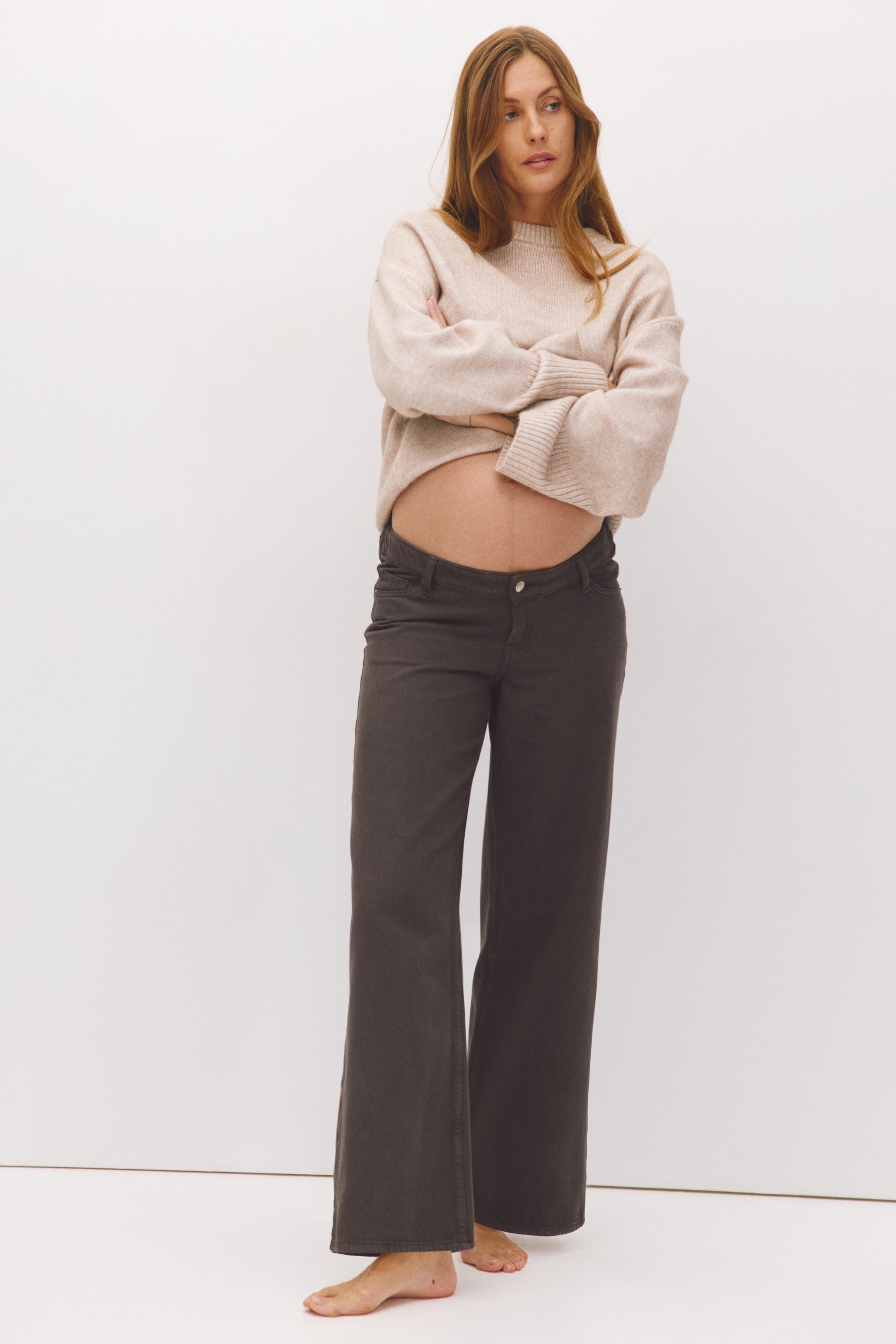 View larger image: These dark grey, wide-leg jeans, featuring a low waist and five-pocket styling, are worn by the model. The jeans are paired with a light beige knit jumper that is cropped or pulled up to show the low-rise fit.