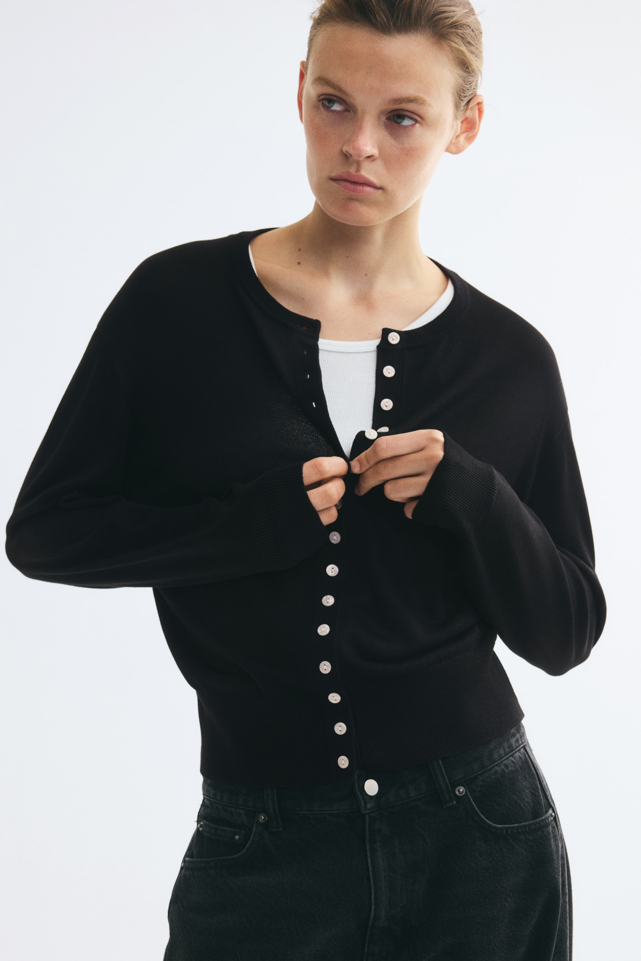 View larger image: A black fine-knit cardigan featuring a round neckline, ribbed trim, and light-coloured front buttons is being worn and adjusted by the model. The cardigan is layered over a white ribbed top and paired with black jeans.