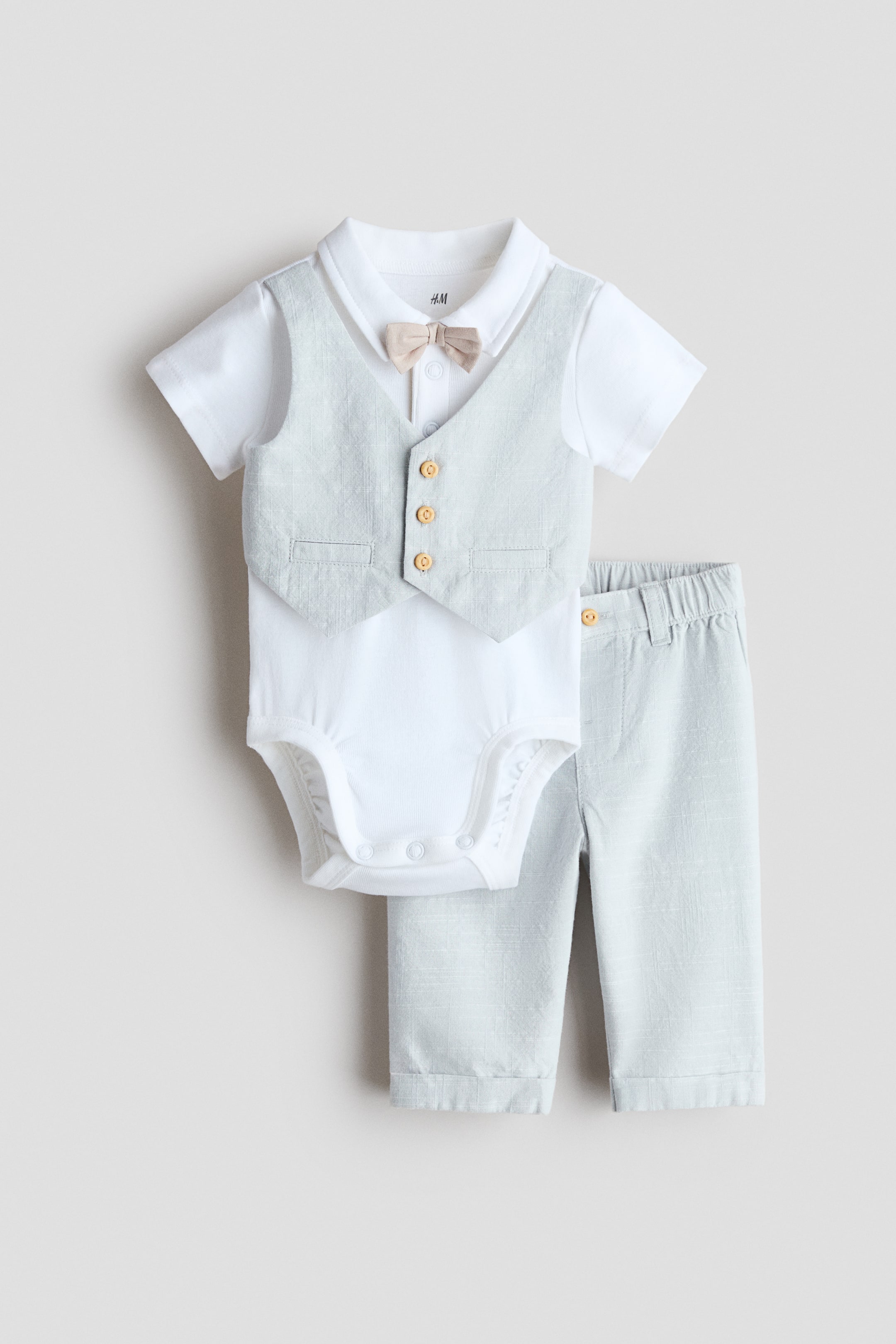 View larger image: A baby's two-piece set is displayed from the front, showcasing a white short-sleeved bodysuit paired with light grey trousers. The bodysuit features a collar, an attached beige bow tie, press-studs at the top and crotch, along with a sewn-on light grey waistcoat that highlights three buttons and two decorative welt pockets. The trousers are designed with an elasticated waist and a buttoned fly.