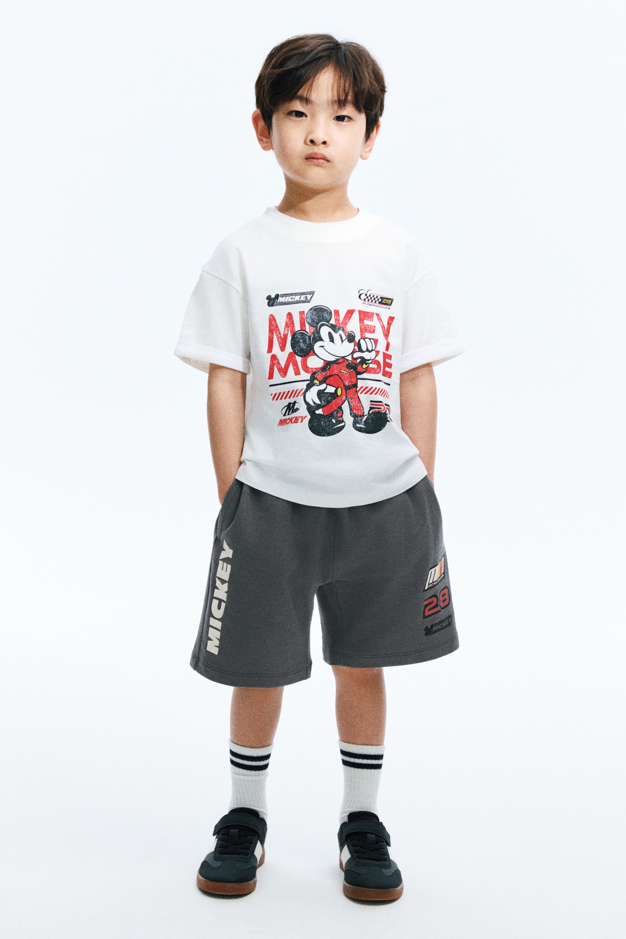 View larger image: A white short-sleeved t-shirt menampilkan cetakan grafik besar Mickey Mouse dalam sut perlumbaan merah, dengan teks "Mickey Mouse" dan lambang perlumbaan. Seluar pendek kelabu gelap melengkapi t-shirt ini, memaparkan logo "MICKEY" menegak di kaki kiri dan grafik "28" dengan jalur berwarna-warni di kaki kanan.