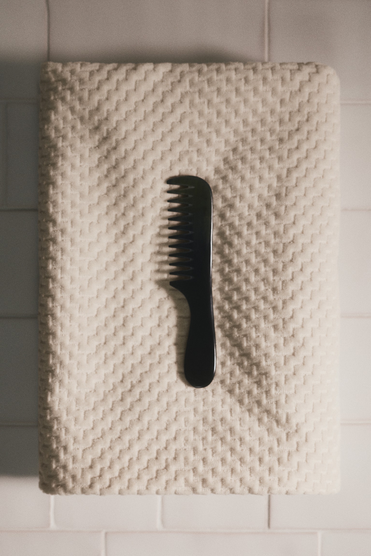 A light beige towel hangs vertically from a dark hook by a fabric loop. It showcases a textured waffle weave pattern and drapes in soft folds. - 1