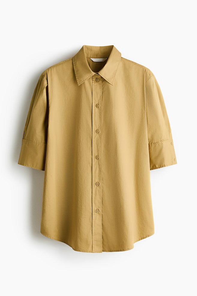 A mustard yellow button-down shirt is shown facing the front, featuring a pointed collar, a central button placket, and elbow-length sleeves with wide, stitched cuffs. The shirt is displaying a relaxed fit and a subtly curved hem. - 1