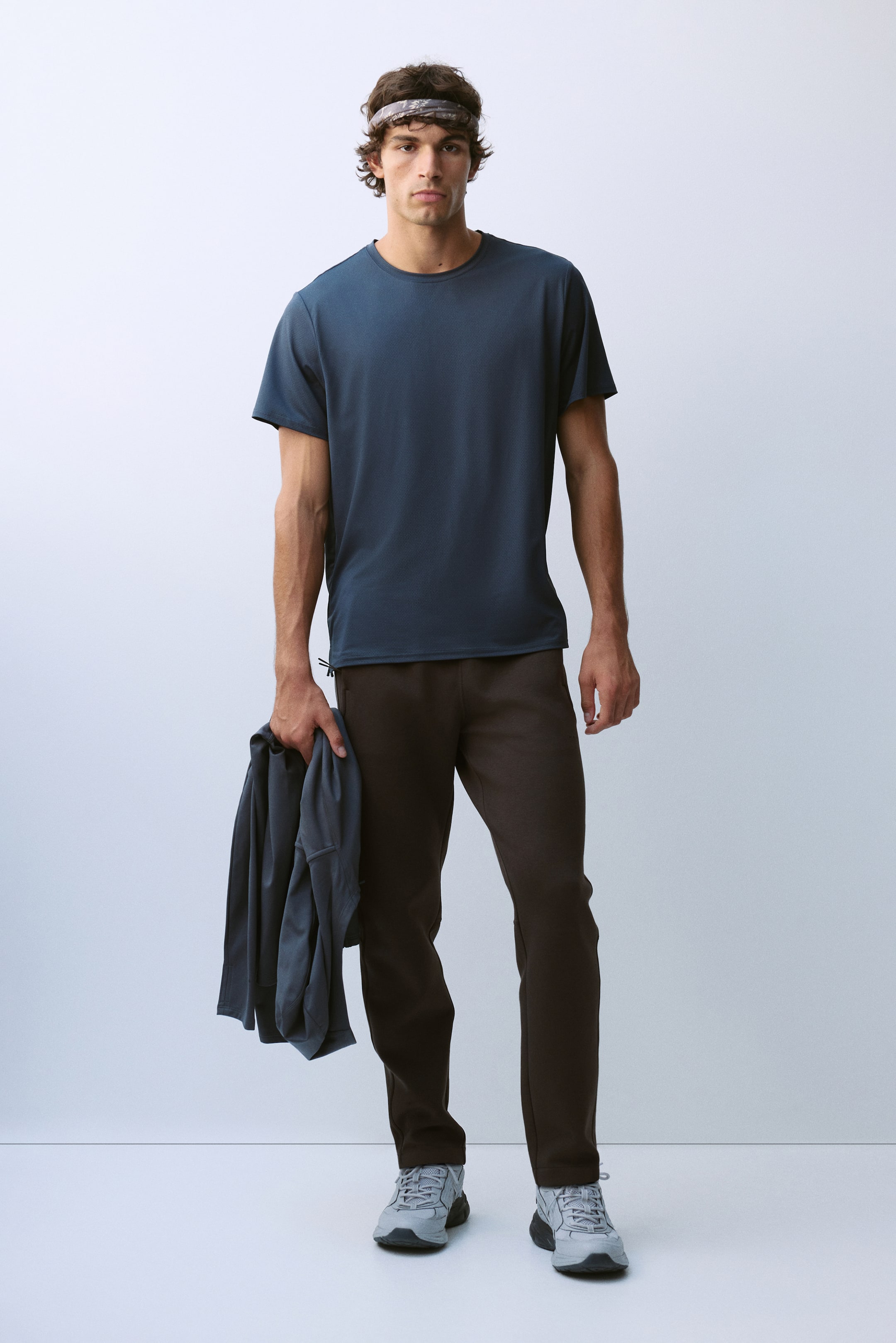 Größeres Bild ansehen: Das Model trägt ein dunkelblaues, kurzärmeliges T-Shirt mit Rundhalsausschnitt und dunkelbraune Hosen. In der rechten Hand des Models wird eine passende dunkelblaue Jacke getragen.
