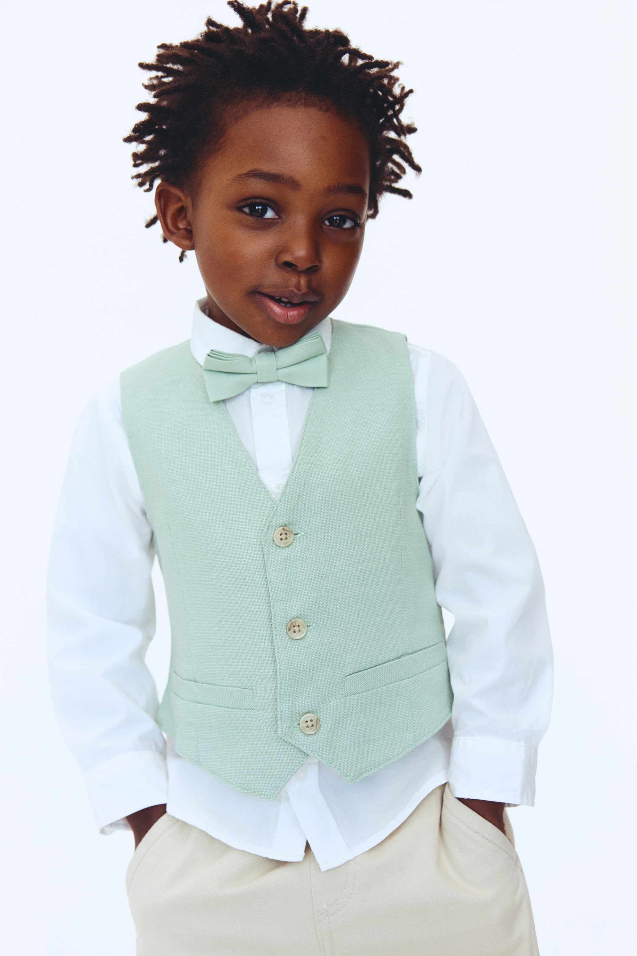View larger image: A light green textured vest paired with a matching bow tie is showcased, styled over a white collared shirt and light beige trousers. The vest boasts a V-neckline, three natural-toned buttons along the front, and two small faux pockets.