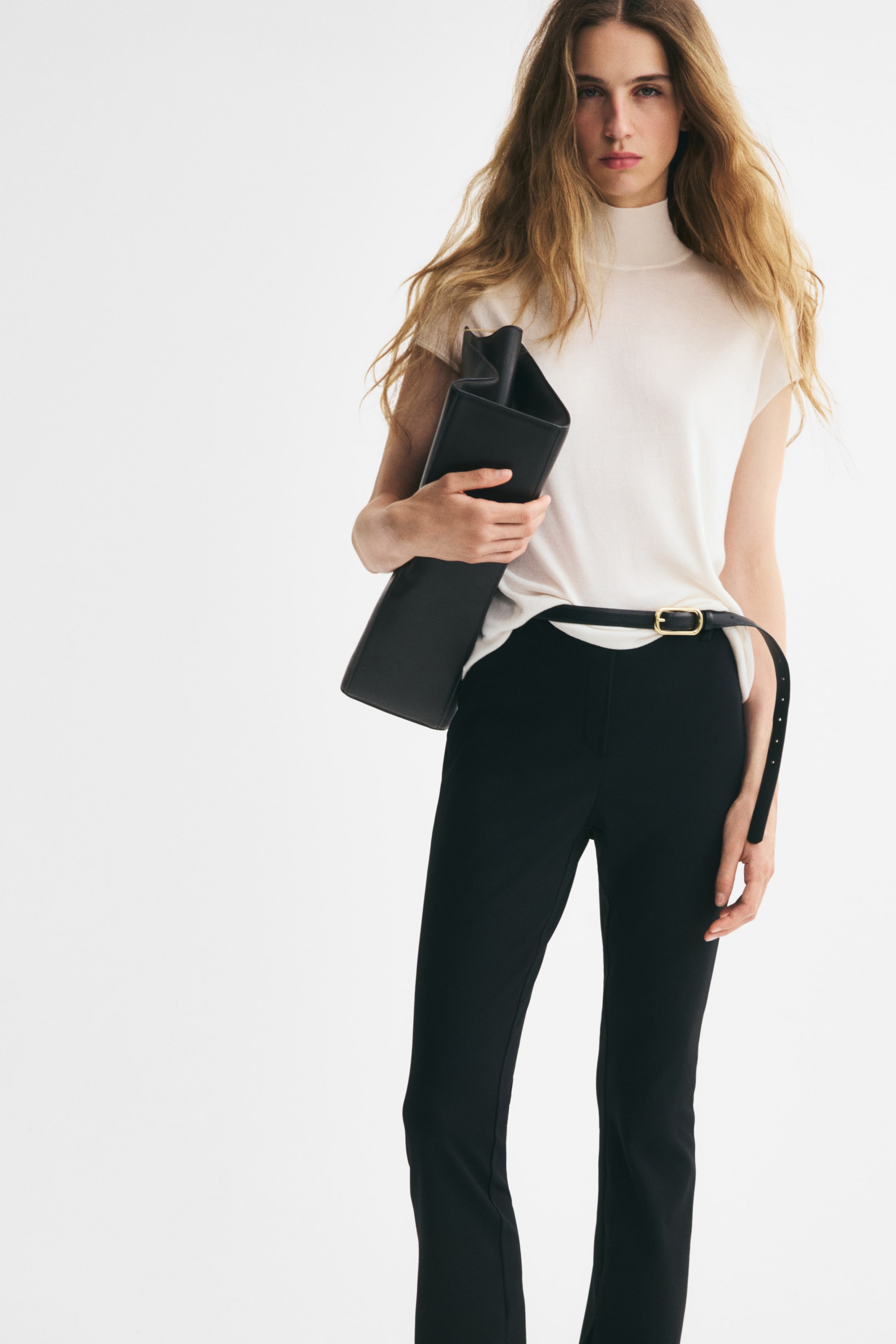 View larger image: A cream-coloured, fine-knit long top is shown from the front, featuring a high neck and cap sleeves. The top is styled with a slim black belt over black trousers, highlighting its soft drape and straight hem.