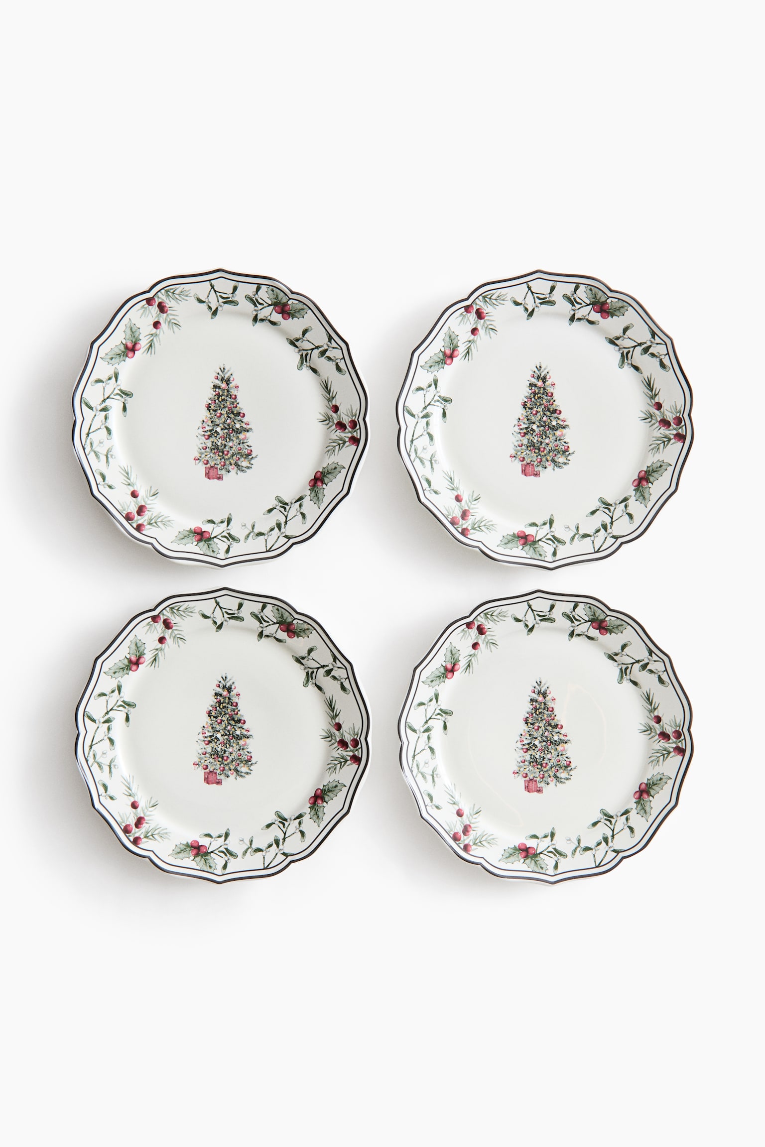 Four white ceramic plates are shown from the front in a two-by-two grid, each featuring a festive design. Each plate has a scalloped edge with a thin dark rim, a central illustration of a decorated evergreen tree, and a surrounding garland of green foliage and red berries. - 1