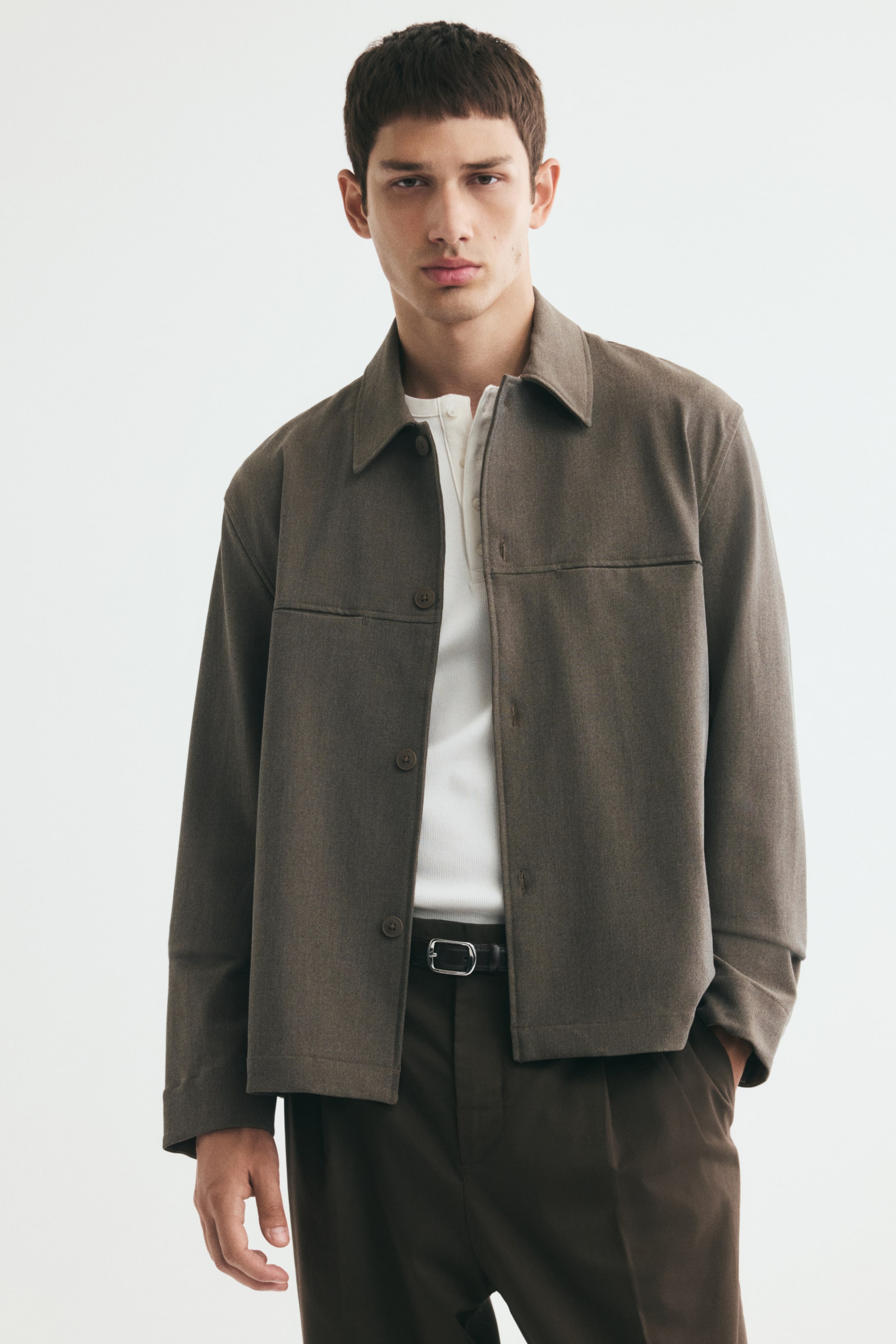 View larger image: A brown overshirt in a soft weave is worn open by the model, featuring a classic collar, front button closures, and a horizontal front yoke with concealed pockets. The overshirt is layered over a white button-neck top tucked into dark brown trousers.