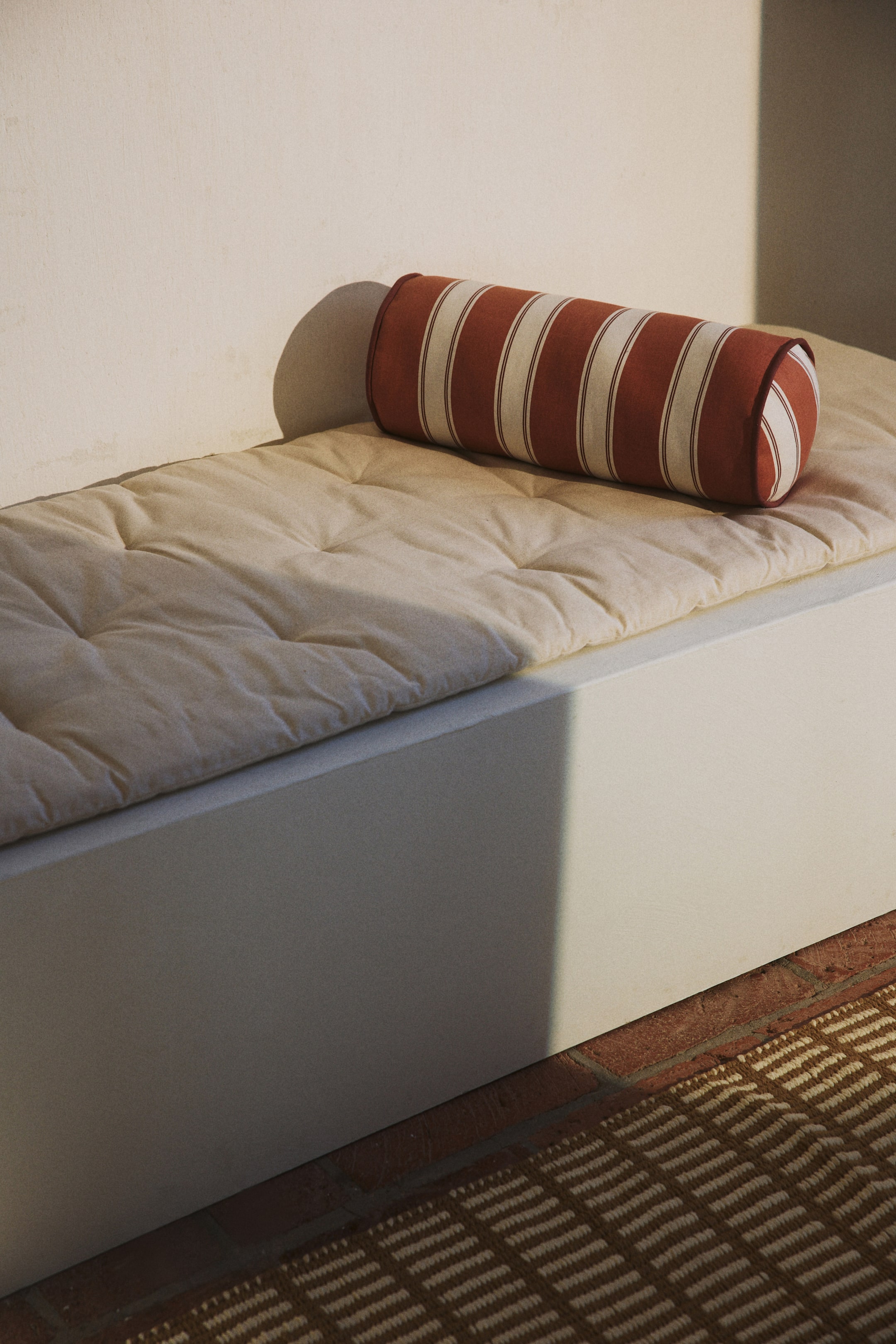 Voir une image plus grande: Un coussin cylindrique repose sur un coussin de banc capitonné, présenté sous un léger angle. Le coussin cylindrique arbore des rayures verticales alternant terre cuite et crème avec des bords passepoilés, tandis que le coussin crème présente plusieurs indentations capitonnées.