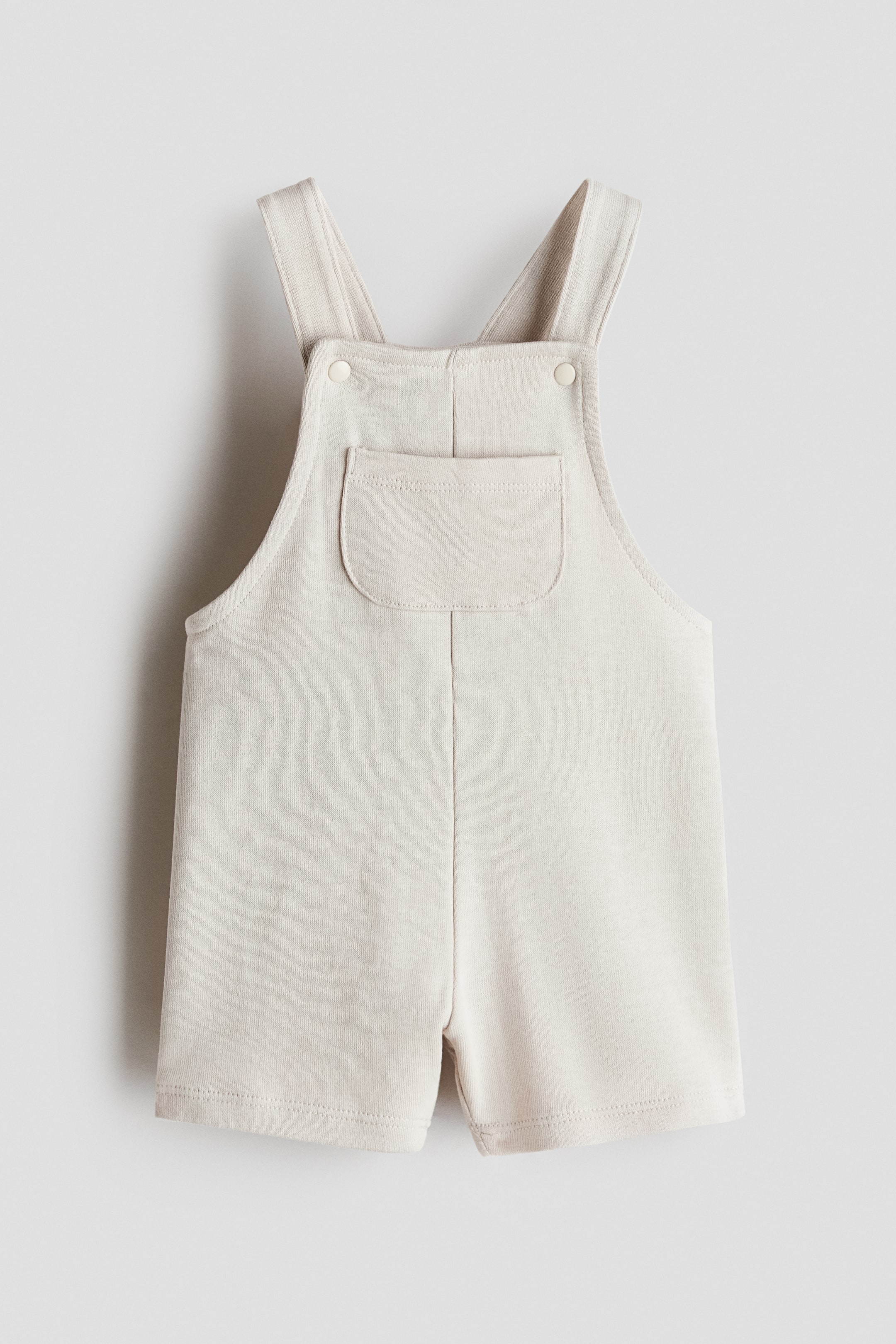 View larger image: Dungaree shorts are showcased front-facing, crafted in a light beige, soft, lightweight sweatshirt fabric. They come with shoulder straps featuring press-stud fastenings, a patch chest pocket, and short legs.