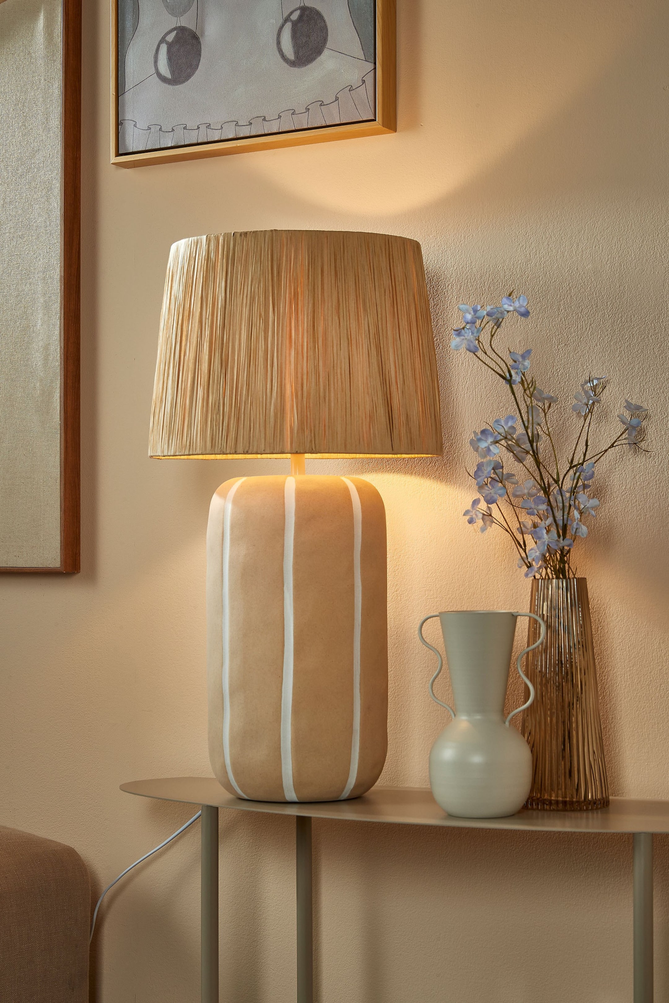 Zobacz większy obraz: Lampa stołowa z kloszem w kształcie stożka wykonanym z naturalnie barwionego plecionego rafii, emitująca ciepłe światło. Jej cylindryczna podstawa jest jasnobrązowa z pionowymi białymi paskami.