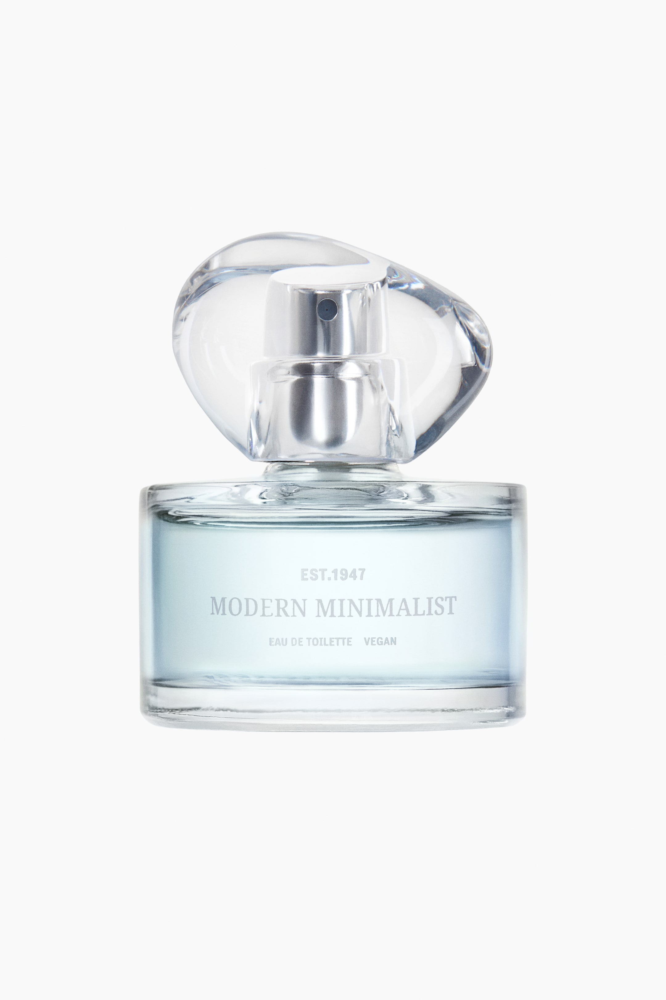 Grotere afbeelding bekijken: Een parfumfles staat rechtop en naar voren gericht, met een heldere rechthoekige glazen basis die lichtblauwe vloeistof bevat. "EST.1947", "MODERN MINIMALIST", "EAU DE TOILETTE" en "VEGAN" worden op de voorkant weergegeven. Een heldere, onregelmatig gevormde dop bedekt een zilveren spraymechanisme.