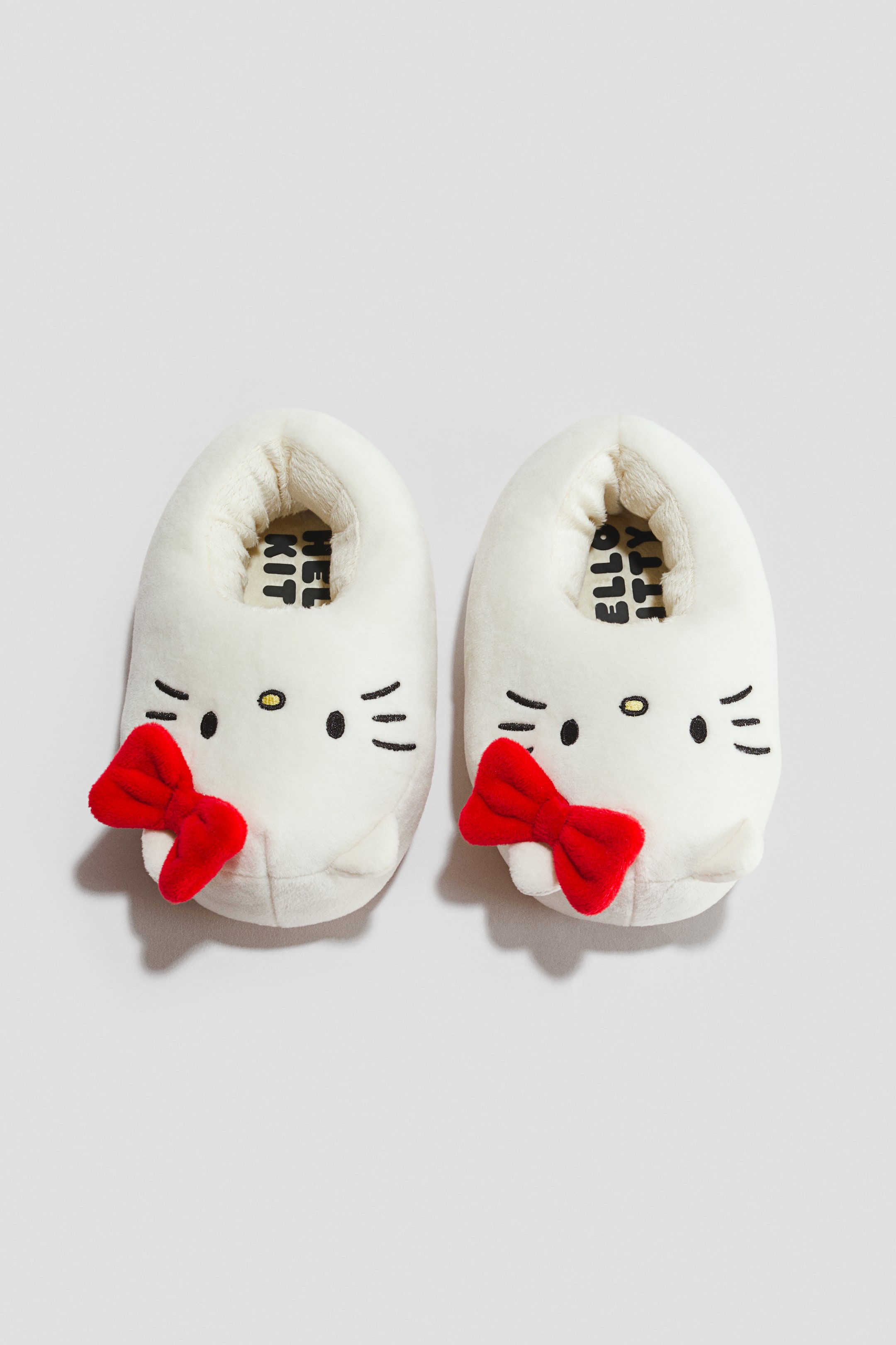 Visa större bild: Ett par vita plyschtofflor med Hello Kitty-motiv har svarta broderade ögon, en gul näsa, morrhår och små rundade öron, med en röd tygrosett som pryder vänster sida av varje tofflas ansikte. Tofflorna är placerade sida vid sida, vända framåt, och visar texten "HELLO KITTY" tryckt i svart på deras innersulor.