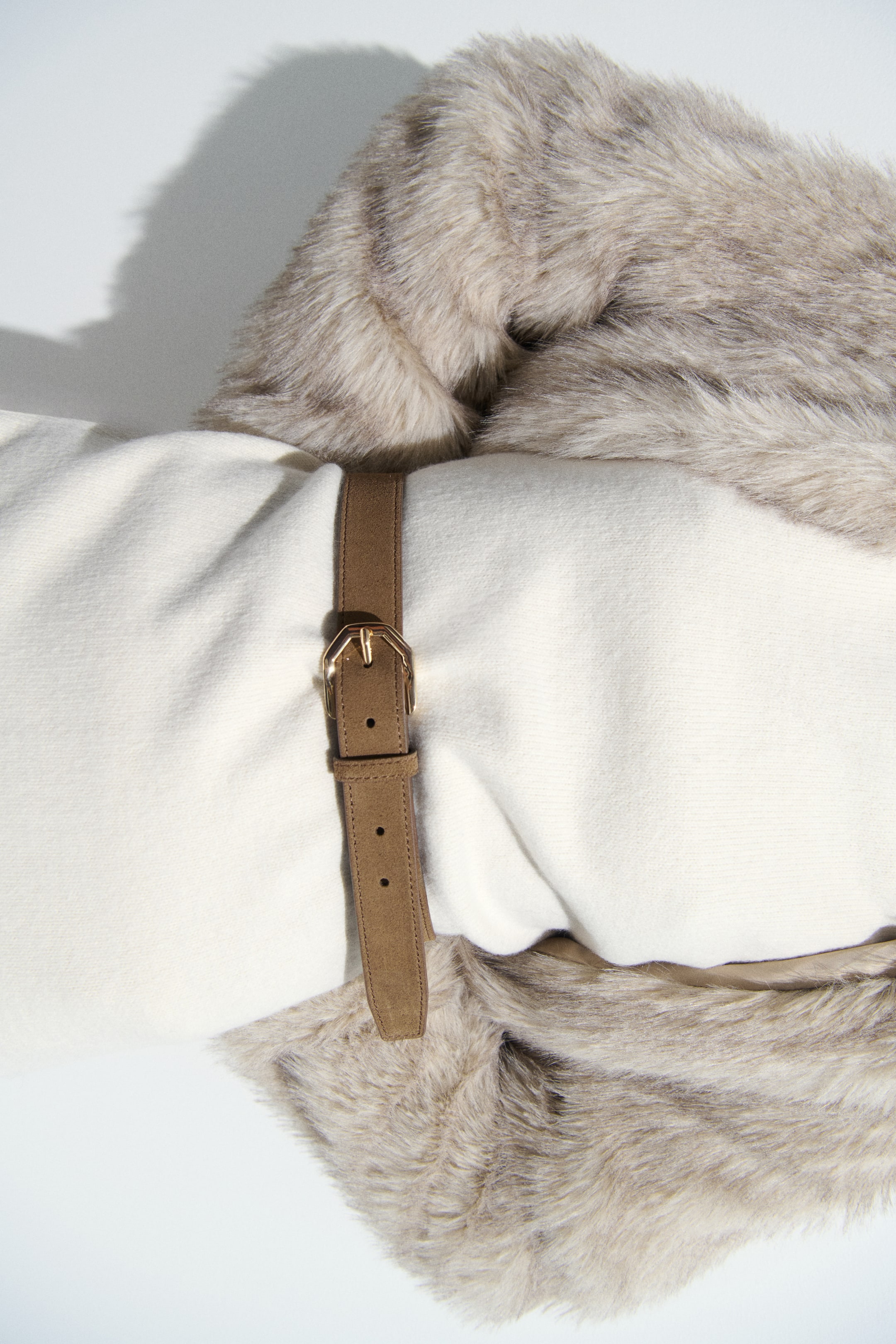 View larger image: A cream-coloured blanket is rolled horizontally, with a light-brown faux fur trim along one edge. A brown strap with a gold buckle cinches the centre of the rolled blanket.