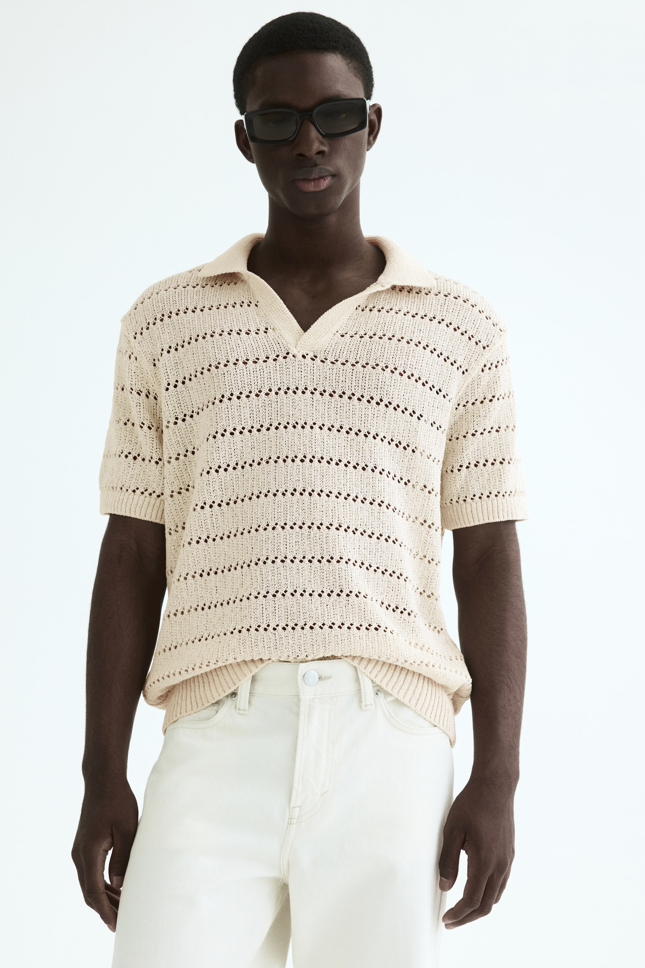 Agrandir l'image: Le polo en maille beige présente des manches courtes avec des poignets côtelés et un ourlet côtelé, complété par un col évasé et une ouverture en V. Des rangées horizontales de détails en maille ajourée sont visibles sur l'ensemble de son corps, créant un design texturé.
