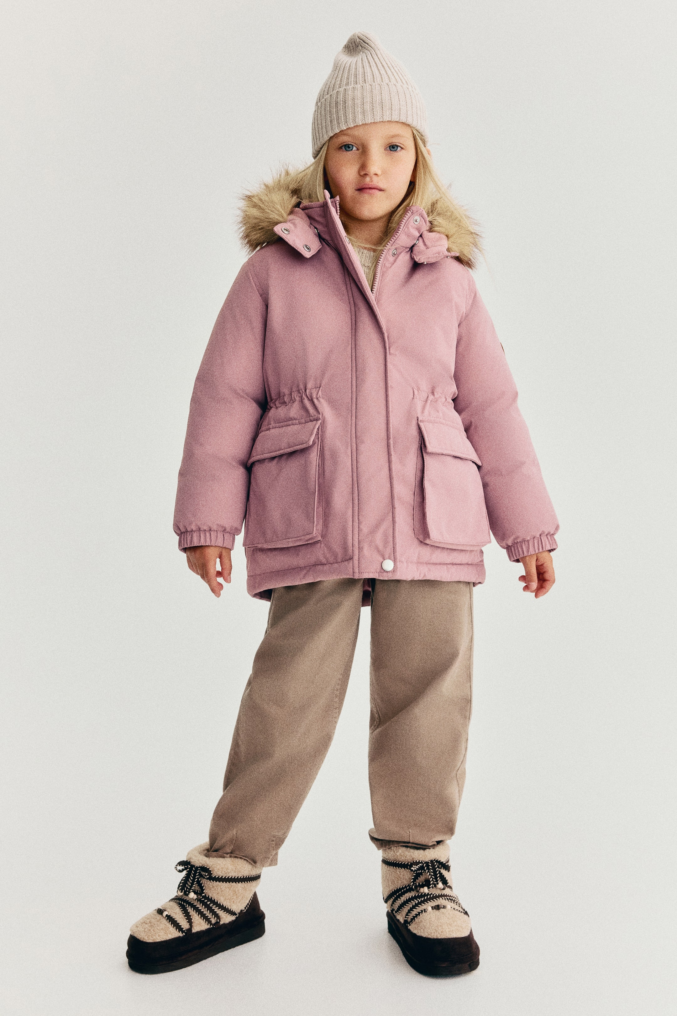 Se større billede: Et barn bærer en lyserød, polstret parka set forfra med hætte med en fluffy faux fur-kant, lynlåslukning foran med vindflap samt store bælgelommer med klapper. Parkaen er designet med elastik i taljen og manchetterne og er stylet her med brune bukser, vinterstøvler og en strikket beanie.