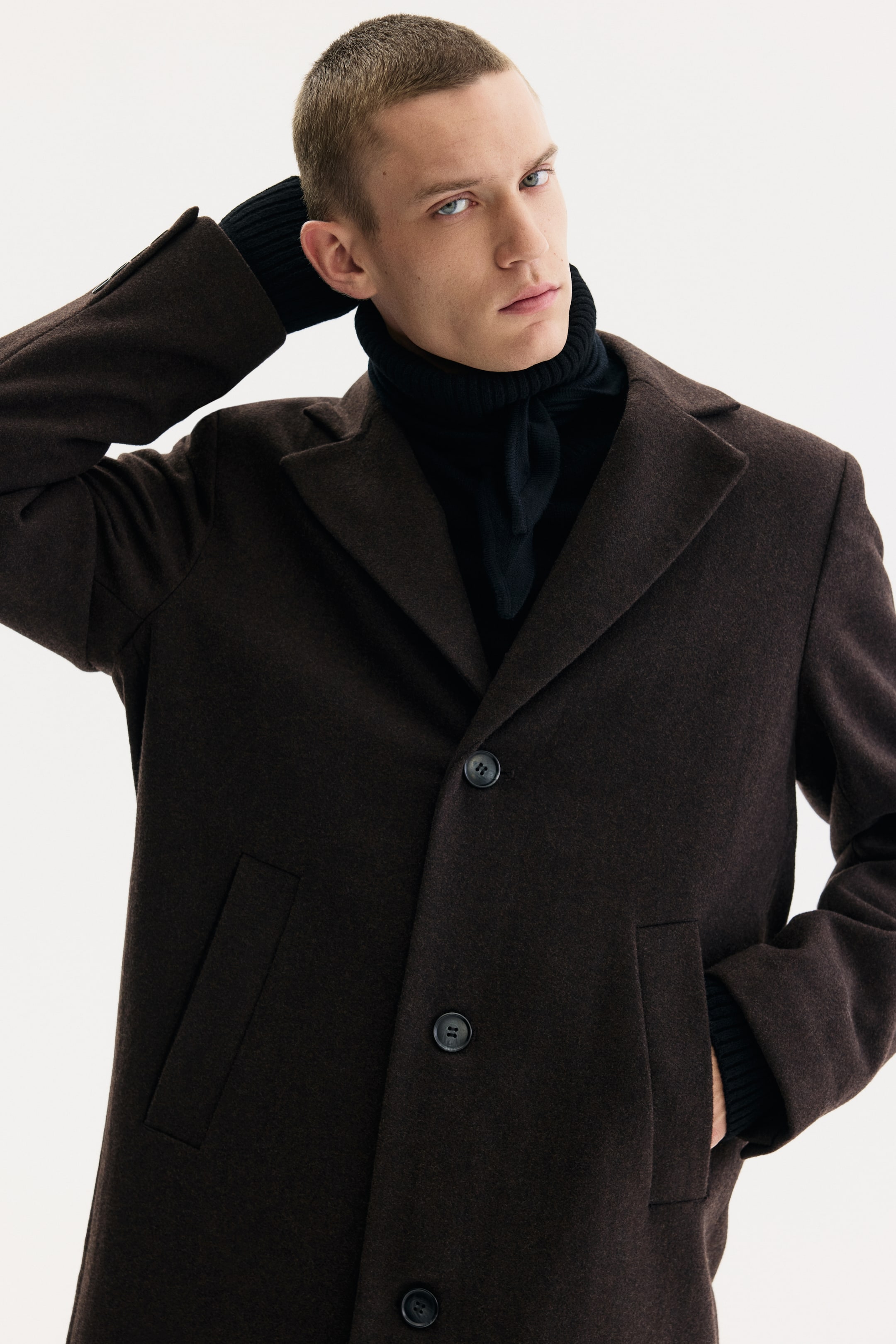 View larger image: A dark brown single-breasted coat is being presented from a front-facing, slightly angled view, featuring notch lapels, three dark buttons, and jetted front pockets, layered over a black ribbed turtleneck with visible cuffs.