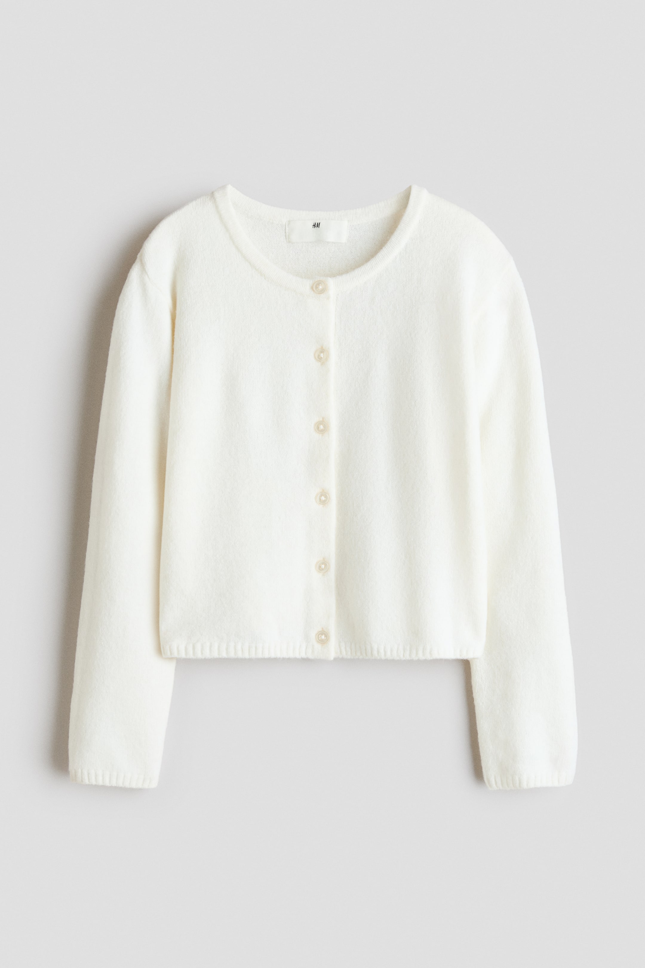 Agrandir l'image: Un cardigan couleur crème est posé à plat, face vers l'avant, avec un col rond, six boutons clairs sur le devant, des manches longues avec des poignets côtelés et un ourlet raccourci côtelé.