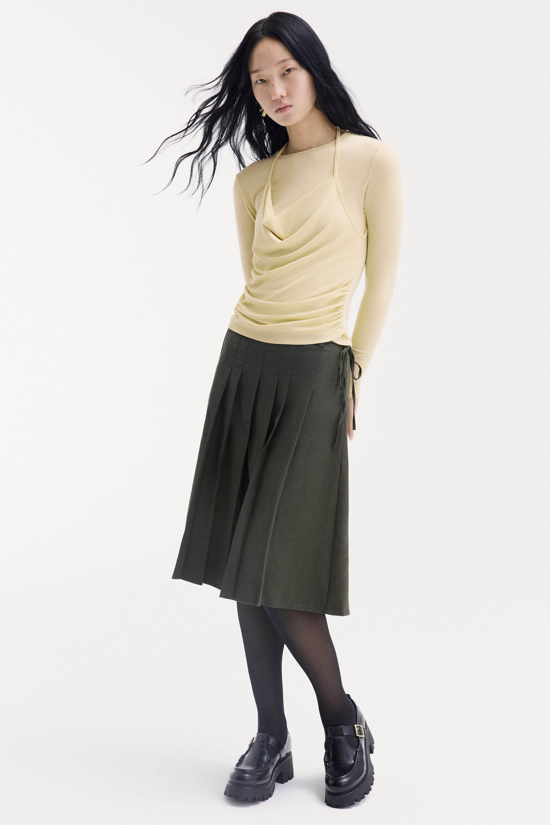 View larger image: The model is wearing a light yellow, long-sleeved jersey top with a high round neckline and a layered cami-style front panel, featuring a draped cowl neck and slim halterneck ties. The top has gathered side seams that create a draped effect at the waist, and is styled with a dark green pleated skirt, black tights, and black loafers.