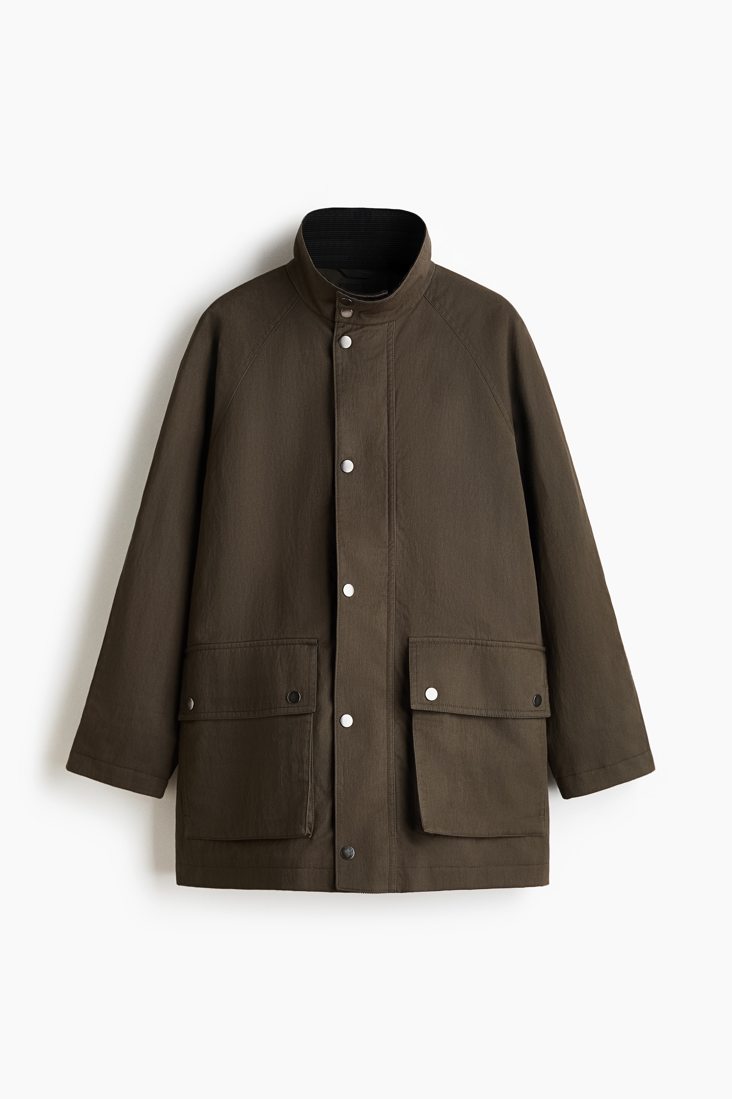 The model is wearing a brown cotton-blend twill parka, featuring a raised stand collar and a partially unzipped front with a press-stud wind flap. The parka has long raglan sleeves and large flap front pockets, and is styled over a dark top and light-coloured trousers.