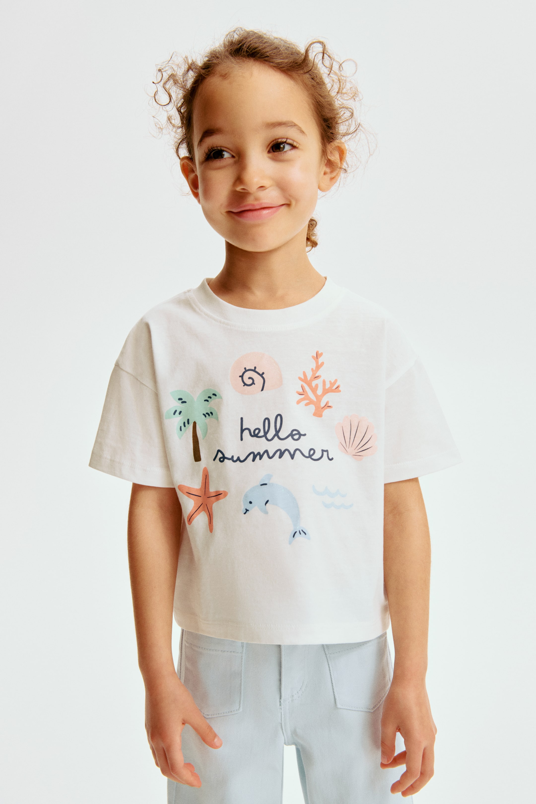 Zobrazit větší obrázek: Bílé tričko s krátkým rukávem zobrazuje na přední straně barevný grafický motiv "hello summer". Grafika zobrazuje palmu, hvězdici, delfína, korál, vlny a různé mušle v jemných, přirozených odstínech.